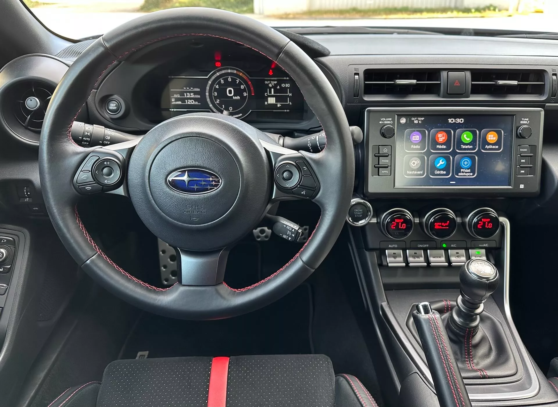 Interior del Subaru BRZ 2023 desde la perspectiva del conductor que incluye un volante deportivo de cuero con controles multifunción y costuras rojas, un cuadro de instrumentos con diales analógicos y una pantalla digital, una consola central con un gran sistema de infoentretenimiento táctil y control climático de zona dual, y un cambio de marchas manual con funda de cuero y detalles en rojo, todo contra un interior negro con acentos deportivos en rojo, destacando las características avanzadas del vehículo y la tecnología.