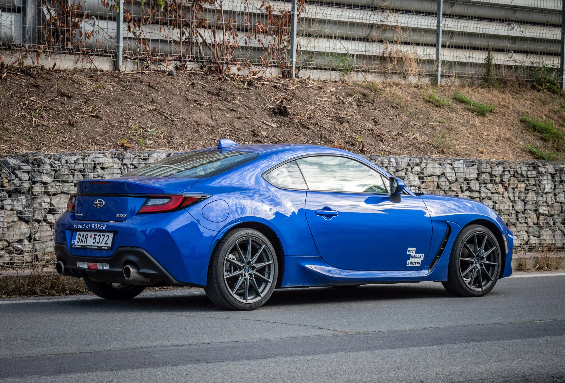 Cupé deportivo Subaru BRZ 2023 de color azul brillante con diseño aerodinámico estacionado en una calle, destacando el arco de la rueda trasera, luces traseras, curvatura de la ventana trasera hasta la tapa del maletero, líneas esculpidas, línea de techo inclinada, ruedas de aleación de múltiples radios, frenos de alto rendimiento, spoiler trasero de bajo perfil, doble tubo de escape en difusor trasero, espejos laterales del color de la carrocería, y un capó largo y deportivo.
