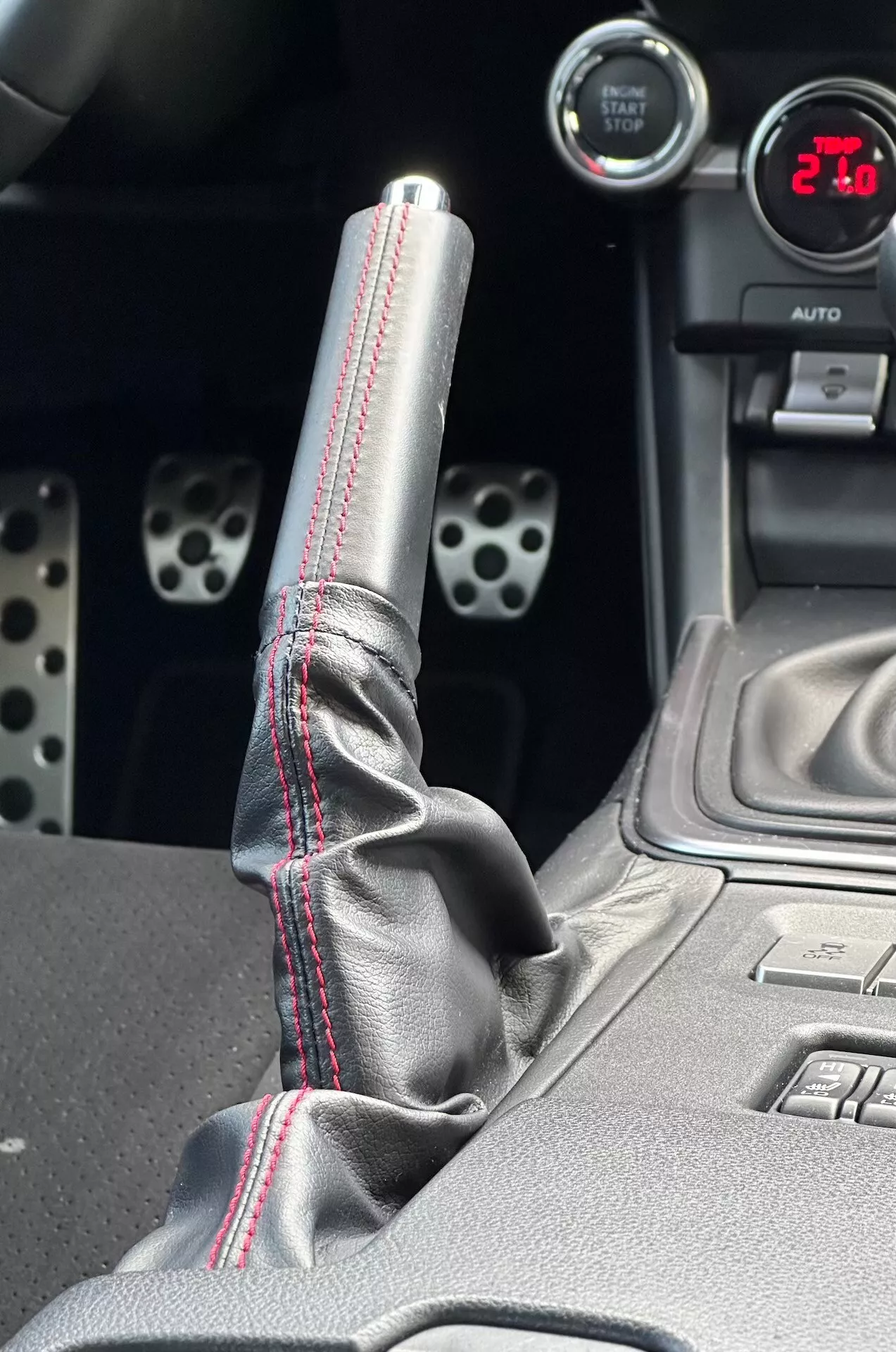 Primer plano del centro de consola del Subaru BRZ 2023 destacando la palanca de cambios metálica con costuras rojas en contraste en la bota de cuero, indicador digital mostrando '21', pedales deportivos de aluminio con agujeros perforados y características tecnológicas como botones de control de estabilidad electrónica.
