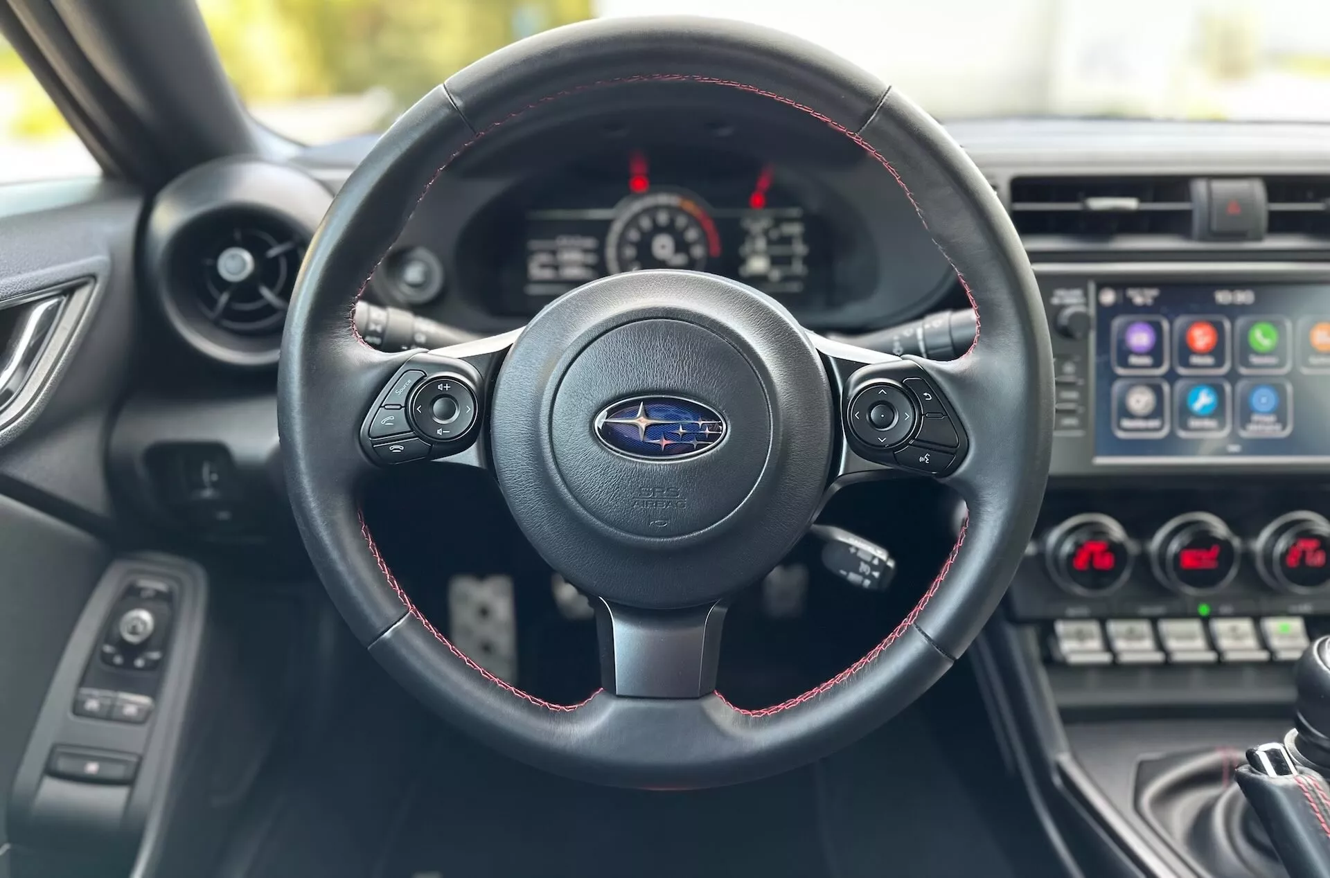 Vista interior del Subaru BRZ 2023 desde el asiento del conductor enfocándose en el volante de tres radios con costuras rojas, logo de Subaru y el tablero de instrumentos con el panel central