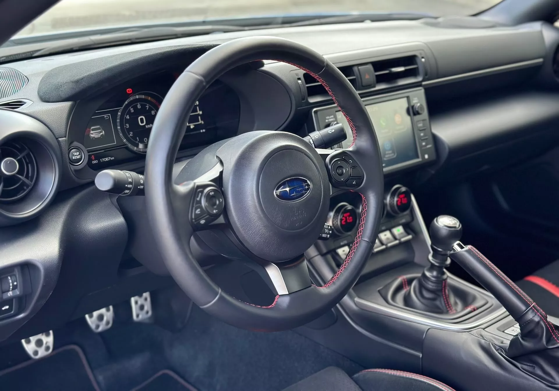 Interior del Subaru BRZ 2023 centrado en el tablero y el cockpit del lado del conductor con vista detallada del volante, el cuadro de instrumentos, la consola central con controles de audio y clima, y la palanca de cambios manual con costuras deportivas rojas