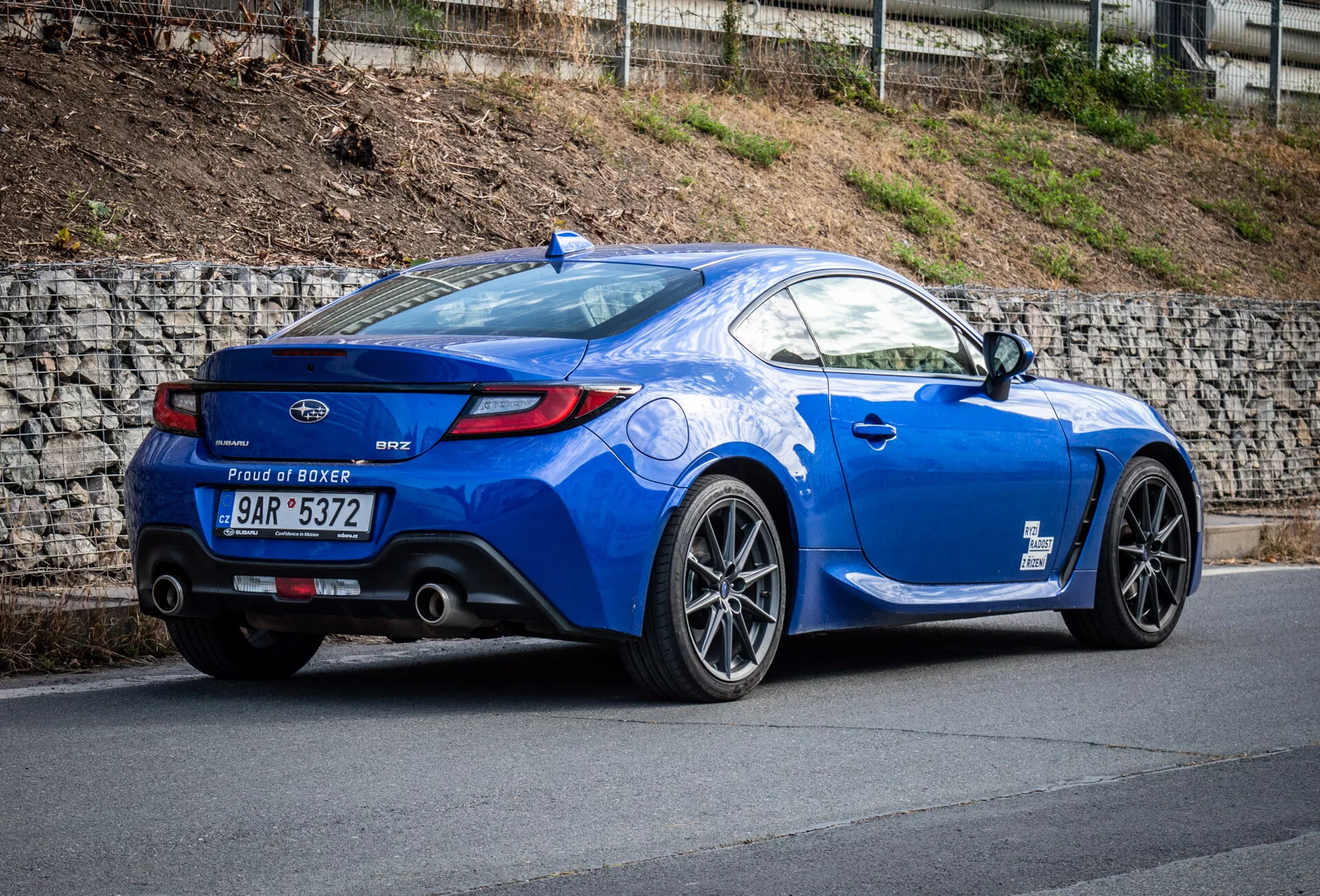 Coupé deportivo Subaru BRZ 2023 en azul metálico estacionado en una pendiente, mostrando su vista trasera de tres cuartos enfatizando el perfil lateral del vehículo, la curvatura trasera, las ruedas de aleación deportivas, las puntas de escape dobles, el alerón labial y la insignia del motor 'Proud of Boxer'.