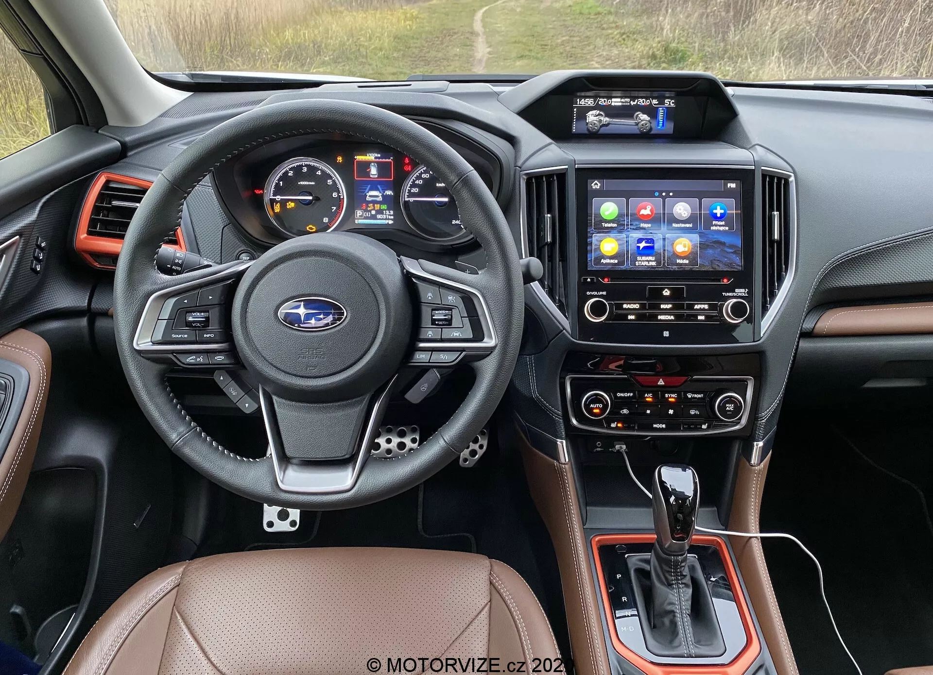 Imagen del interior desde la perspectiva del asiento del conductor de un Subaru Forester 2020, enfocada en el tablero, volante, sistema de infoentretenimiento y consola central. El volante de cuero de tres radios incluye controles multifuncionales para audio, teléfono, comandos de voz y control de crucero, con el emblema de Subaru centrado. El panel de instrumentos incluye una pantalla digital y medidores analógicos de velocidad y RPM. La consola central alberga la palanca de cambios de transmisión automática, controles adicionales y posiblemente un selector de modo de conducción. La pantalla táctil de infoentretenimiento con varios íconos de aplicaciones ofrece navegación, medios, conectividad telefónica y configuraciones del vehículo. Abajo, los controles de clima sugieren ajustes de doble zona para el conductor y el pasajero delantero. Los acabados interiores incluyen asientos de cuero o simil, componentes plásticos negros y acentos metálicos. Las salidas de aire flanquean el sistema de infoentretenimiento y los bordes del tablero. Esta imagen se clasifica en interiores de automóviles, tecnología automotriz, tableros, sistemas de infoentretenimiento y controles de clima.