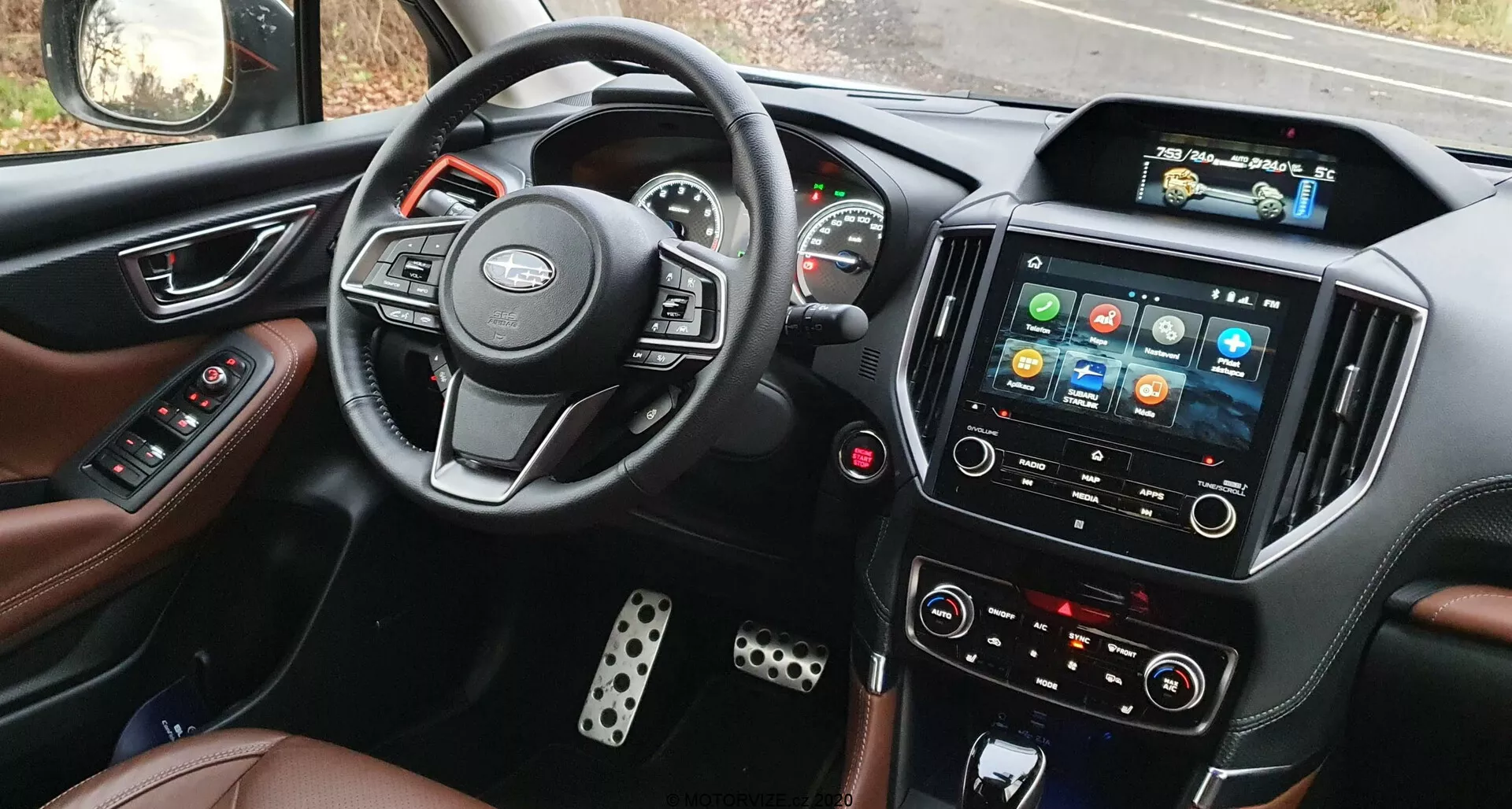 Vista interior del Subaru Forester 2020 desde el asiento del conductor, mostrando el volante con controles integrados, el cuadro de instrumentos parcialmente visible con indicadores analógicos y pantalla digital central, un sistema de infoentretenimiento con pantalla táctil prominente y iconos coloridos, botones de control de clima manuales, asientos con tapicería de cuero y cubrepedales metálicos.