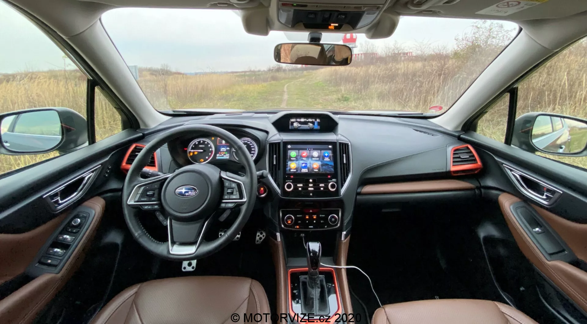 Vista interior de la cabina delantera del Subaru Forester 2020, mostrando un volante multifuncional con controles, un cuadro de instrumentos digital, consola central con sistema de infoentretenimiento con pantalla táctil, palanca de cambios automática y un interior de dos tonos con cuero marrón y detalles metálicos, visible desde el área de asientos traseros hacia el tablero.