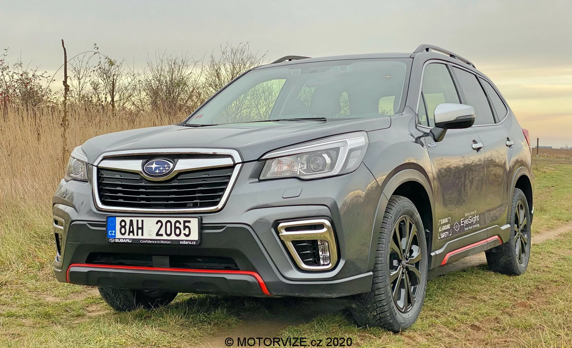 La imagen muestra un SUV Subaru Forester 2020 de color plateado estacionado al aire libre durante el día con luz natural. Capturada desde un ángulo frontal de tres cuartos, la foto muestra claramente el lado izquierdo frontal del vehículo, incluida su parrilla hexagonal con el emblema del fabricante, los elegantes faros LED o HID, las luces diurnas en forma de C alrededor de los faros antiniebla y el parachoques delantero atlético con elementos de placa protectora plateada. El capó esculpido tiene crestas elevadas y los abultamientos de los guardafangos le dan una postura musculosa. El perfil lateral presenta una línea de carácter prominente, revestimientos de plástico negro en la parte inferior de las puertas, llantas de aleación cubiertas de tierra, rieles en el techo y un espejo lateral parcialmente visible con señales de giro integradas. Una insignia en el guardabarros delantero indica un nivel de equipamiento especial o el tipo de motor. El entorno sugiere un uso ligero fuera de carretera, enfatizado por las salpicaduras de barro en los lados del vehículo.
