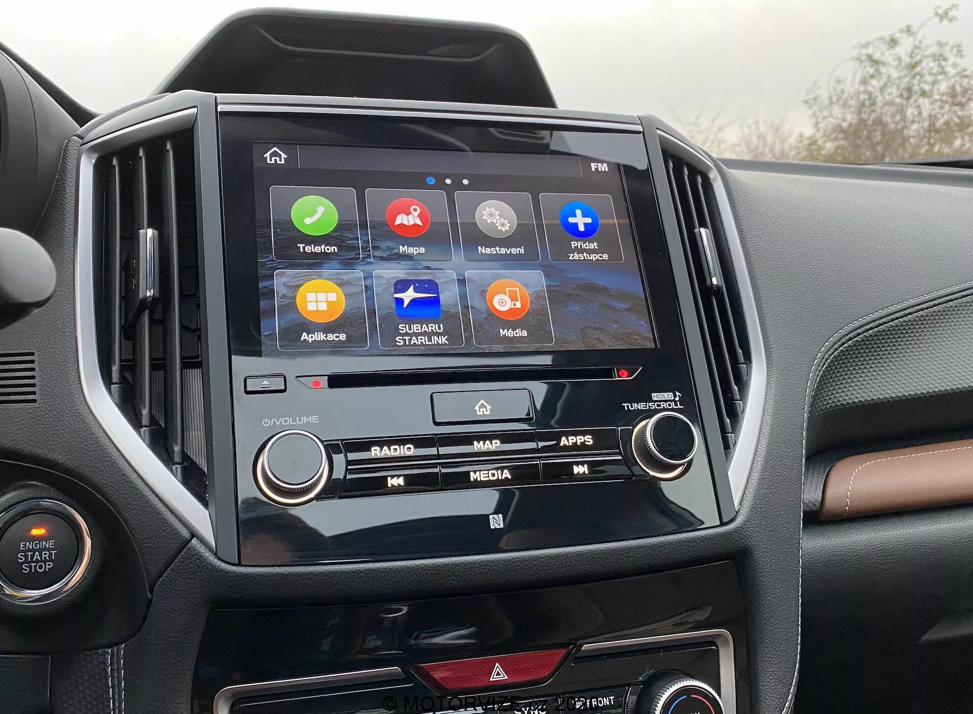 La imagen muestra la consola central de un Subaru Forester 2020, destacando su sistema de infoentretenimiento y el interior. En el centro de la imagen se encuentra la pantalla táctil con iconos para 'Teléfono', 'Mapas', 'Reproduciendo ahora', 'Mensajes', 'Subaru Starlink', 'Medios' y 'Aplicaciones', indicando diversas funcionalidades como conectividad, navegación y entretenimiento. La pantalla de infoentretenimiento está enmarcada en un bisel negro brillante que contrasta con el tablero bicolor. También son visibles los botones físicos y los mandos de control del clima, ilustrando una combinación de controles digitales y manuales.