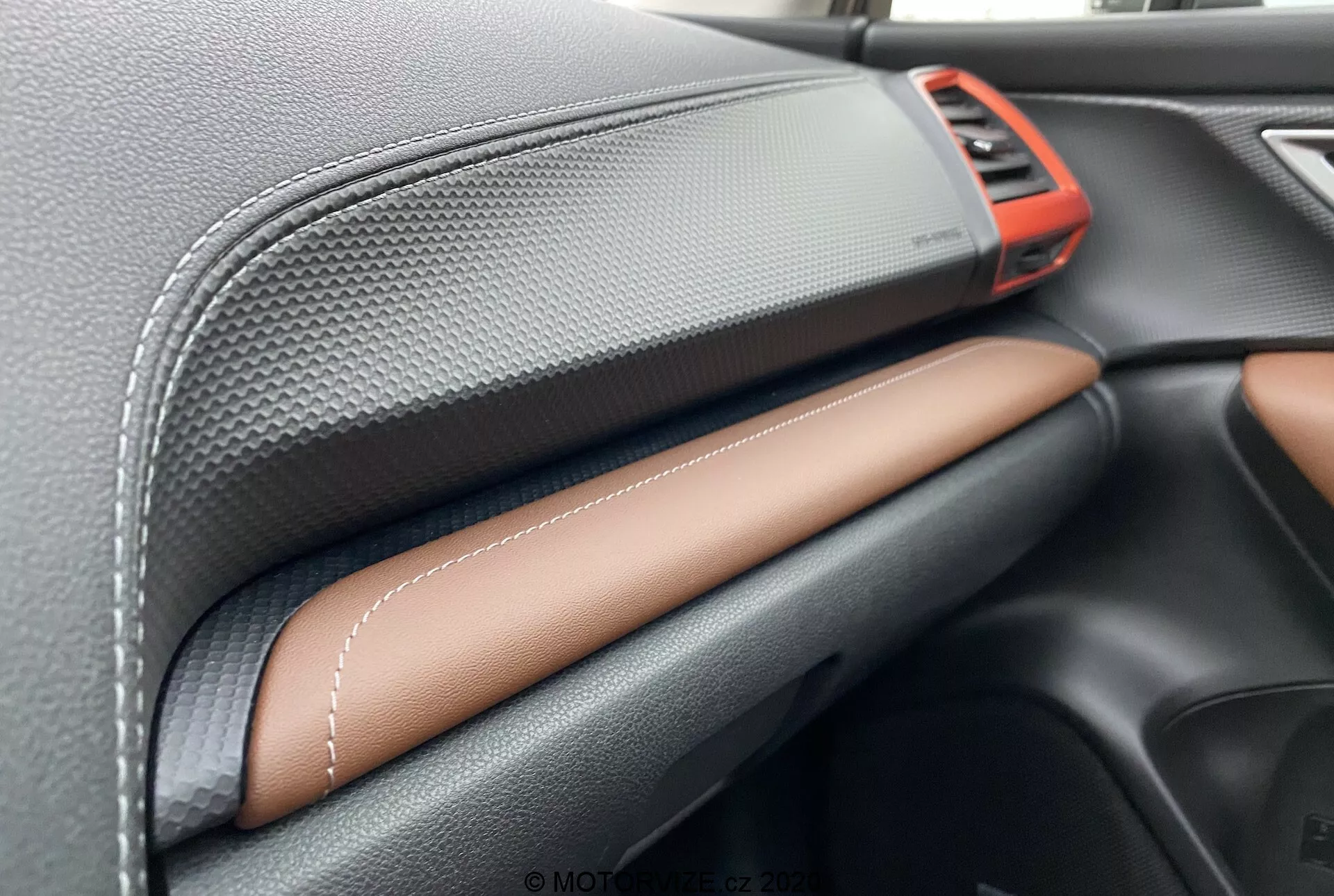 Vista de cerca del interior del Subaru Forester 2020 centrada en el tablero. Incluye una rejilla de ventilación metálica roja brillante en la parte superior derecha, material negro texturizado como fibra de carbono en la parte superior del tablero, cuero sintético con costuras en contraste en color marrón claro o beige en el centro y un elegante compartimento para guantes negro debajo. Esto muestra la artesanía y la calidad de los materiales premium.