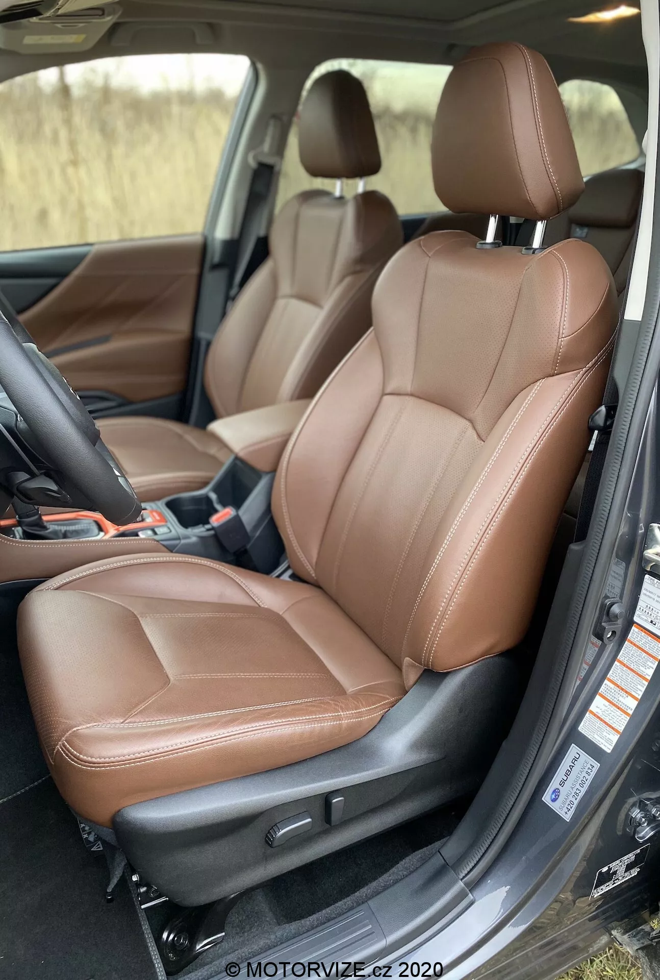 Esta imagen captura el lujoso interior de un Subaru Forester 2020, enfocándose en el asiento del pasajero delantero, que también podría fungir como el asiento del conductor en una configuración de manejo a la izquierda. Tomada desde un ángulo ligeramente elevado, la imagen proporciona una vista clara del asiento de cuero marrón con elementos de diseño ergonómico, incluidos refuerzos laterales y soporte contorneado. Se pueden ver características como el reposacabezas ajustable, posibles opciones integradas de calefacción/refrigeración/masaje y funciones ajustables eléctricamente demostradas por botones y una palanca de control similar a un joystick. La puerta está abierta, revelando parte del entorno exterior, y el panel interior de la puerta se alinea armoniosamente con el diseño del asiento. La alfombrilla de moqueta y la robusta construcción sugieren la alta calidad del vehículo del segmento de lujo, mejorando la comodidad y la tecnología avanzada en el área del asiento.