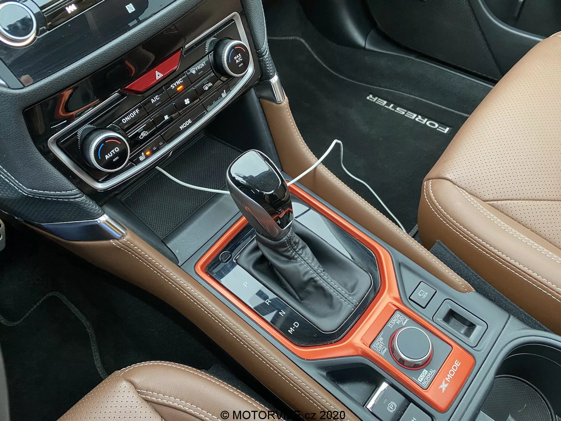 Vista interior del Subaru Forester 2020 centrada en la consola central desde una perspectiva aérea, con una moderna palanca de cambios automática, panel negro con detalles en rojo anaranjado, interfaz de control con botones y diales, botones y pantalla de control climático, botón de arranque/parada sin llave, selector de modos de conducción, tapicería de cuero color avellana con costuras en contraste, 'BOSE' en relieve en el cuero, y vista parcial del asiento del pasajero y el reposabrazos, destacando la mezcla de deportividad y lujo del vehículo.
