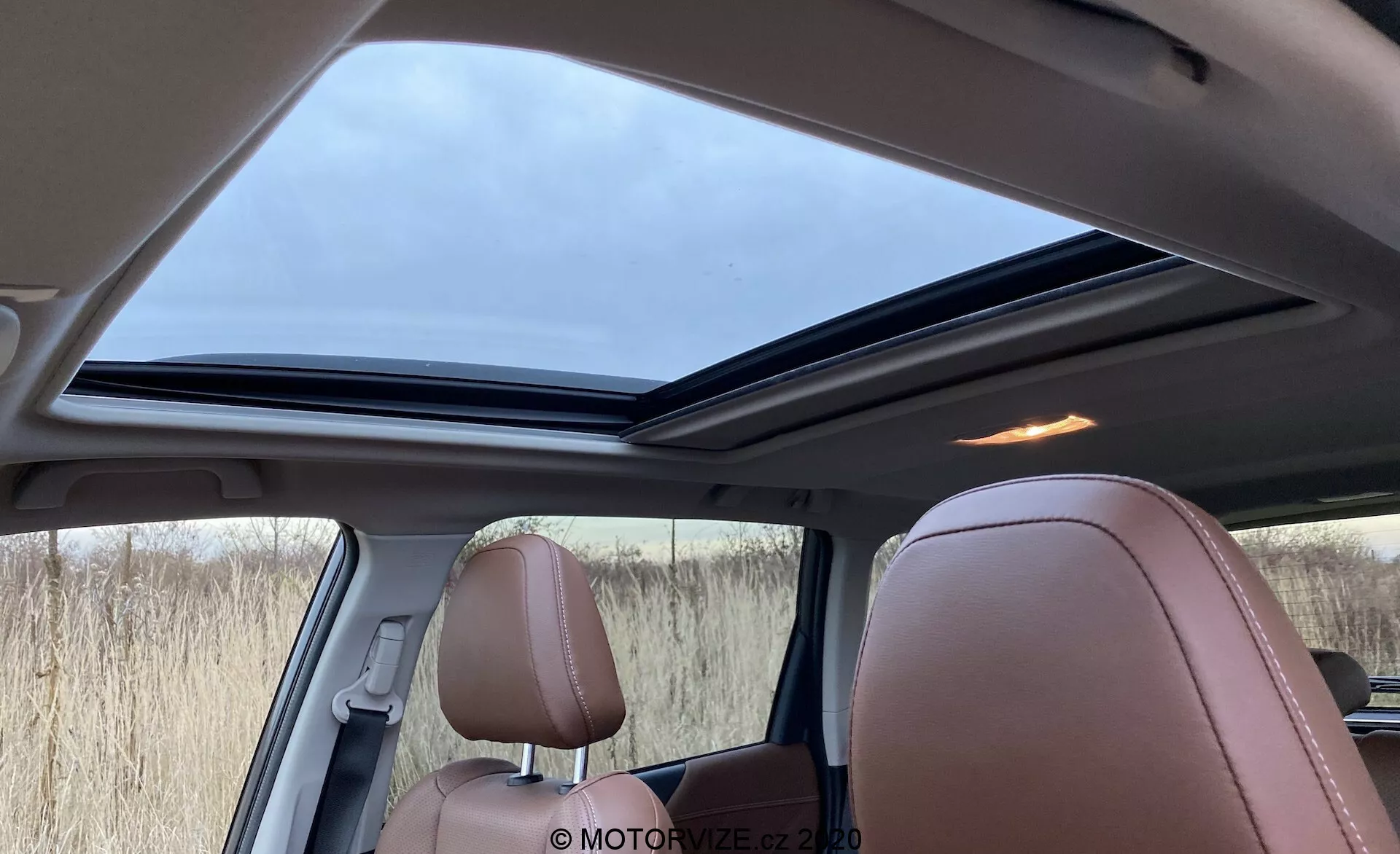 Vista interior del Subaru Forester 2020 desde la perspectiva del pasajero trasero, destacando el gran techo solar panorámico en una posición ligeramente abierta, los lujosos asientos de cuero marrón claro con costuras detalladas y una combinación de acabado interior en marrón, negro y beige. También visibles son una rejilla de altavoz cerca de un pilar y ventanas laterales tintadas, iluminadas por la luz suave del día a través del cielo nublado.