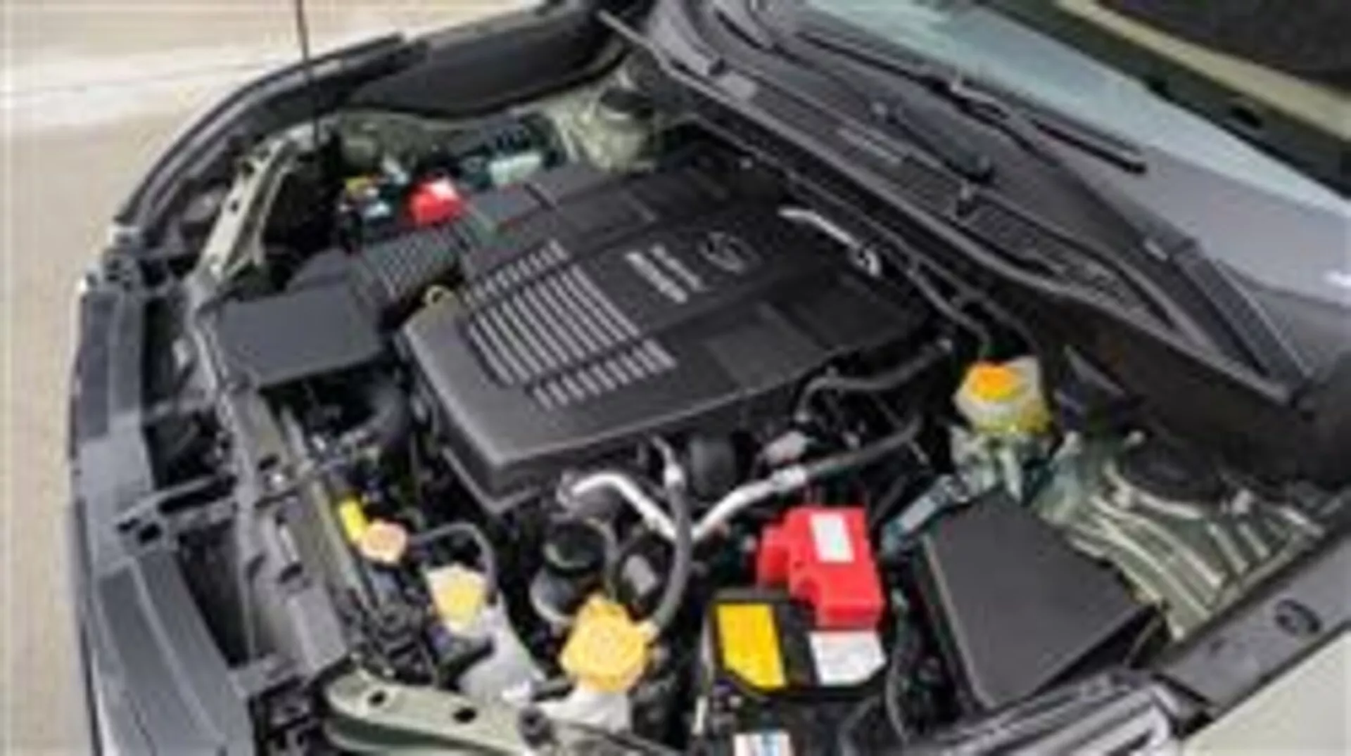 Vista de cerca del compartimiento del motor de Subaru Forester 2015 mostrando la batería, los depósitos de líquido, la caja del filtro de aire, tuberías y mangueras variadas, partes mecánicas y la cubierta del motor