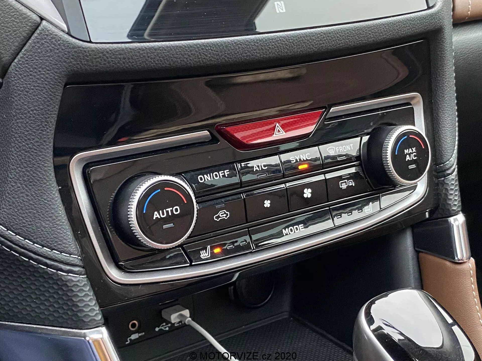Vista detallada de la consola central de un Subaru Forester 2020, enfocándose en el panel de control del clima. Son visibles los controles de climatización de doble zona con dos grandes perillas para el ajuste de la temperatura, botones para las luces de emergencia, SYNC, MAX AC y desempañador del parabrisas delantero, así como un panel de pantalla LED (no iluminado). A continuación, hay controles adicionales para la dirección del flujo de aire, velocidad del ventilador, recirculación, desempañador trasero y encendido/apagado del aire acondicionado, todo en un acabado que asemeja a fibra de carbono. También se vislumbra parte de la palanca de cambios y un portavasos/compartimento de almacenamiento.