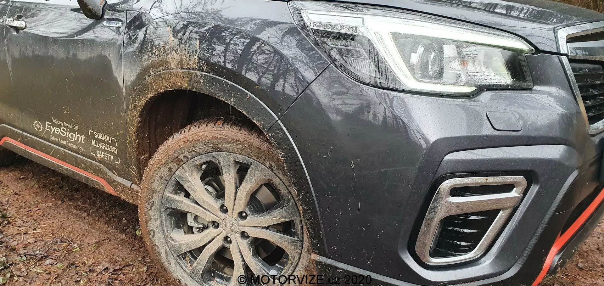 Vista del cuarto delantero izquierdo de un Subaru Forester 2020 embarrado, capturado desde un ángulo bajo, destacando su despeje del suelo, llanta de aleación multirradio intrincada, diseño del parachoques inferior y recortes angulares de la parrilla. El vehículo de color oscuro, probablemente negro o gris oscuro, muestra signos de uso todoterreno con salpicaduras de barro en la rueda delantera, el faldón lateral y el estribo. Las características notables incluyen un gancho de remolque integrado en el parachoques, un protector de bajos metálico delineado en rojo para protección inferior y una mención 'EyeSight', que sugiere capacidades avanzadas de asistencia al conductor.