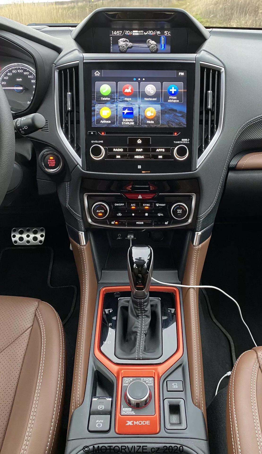 La imagen captura la consola central interior de un Subaru Forester 2020 desde la perspectiva del conductor, enfocándose en la pila y túnel centrales. Una pantalla táctil de alta resolución y orientación vertical domina la sección superior, mostrando un colorido menú principal con iconos para navegación, teléfono, medios y ajustes del automóvil. Debajo de la pantalla, se pueden ver dos salidas de aire con acentos cromados y controles táctiles para un sistema de clima de doble zona con funciones avanzadas, incluyendo botones de desescarche. La sección inferior destaca un botón de encendido/apagado, una palanca de cambios automática con acabados en cuero y cromo, selectores de modos de conducción como 'X-MODE' y varios botones para configuraciones de manejo. Un freno de estacionamiento electrónico, función de retención automática y materiales lujosos como cuero con costuras de contraste, metálico y acabados de plástico brillante mejoran la sensación premium. La toma enfatiza la tecnología avanzada del vehículo, comodidad, conectividad y adaptabilidad.