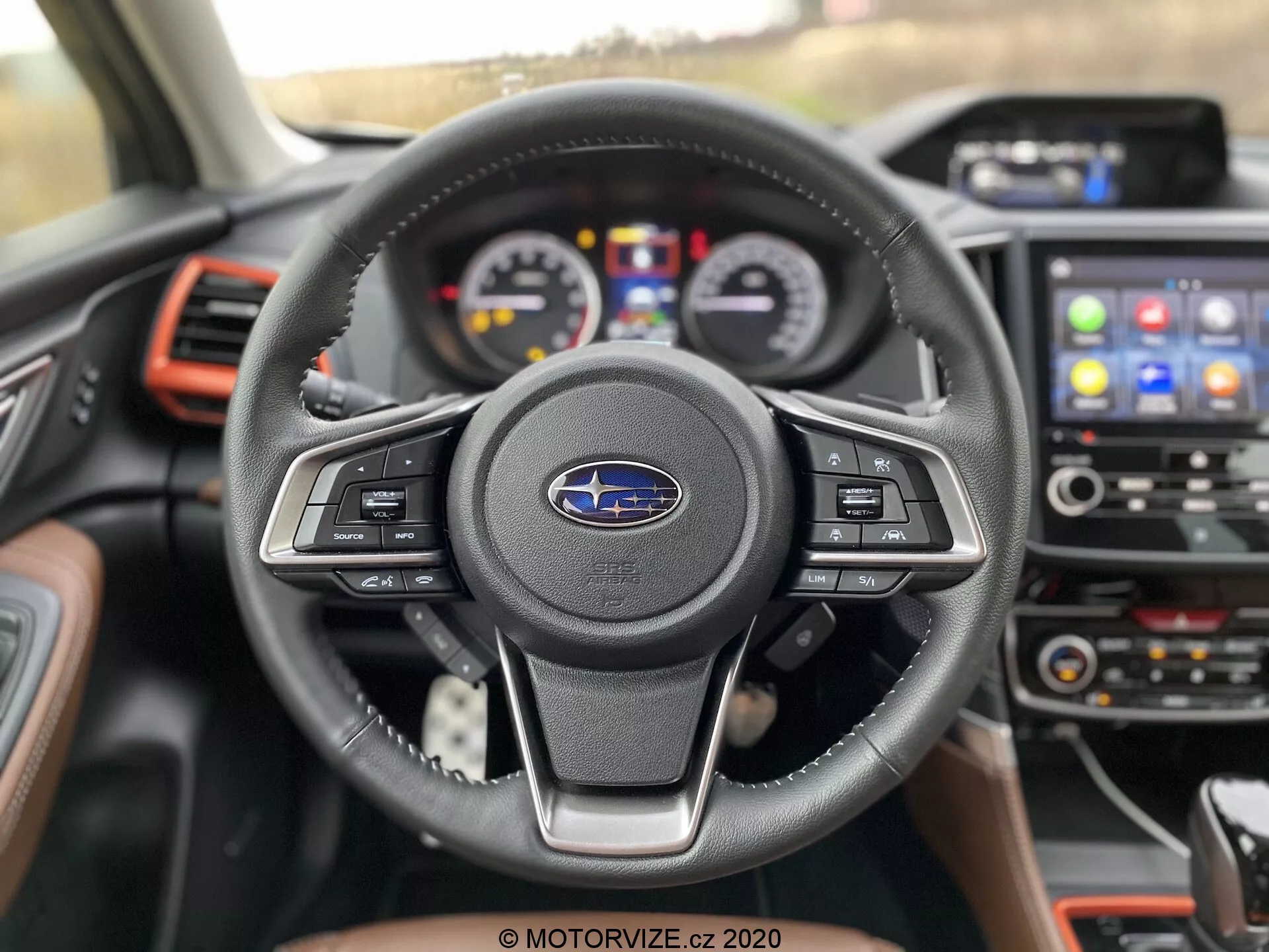 Vista de primer plano del interior del Subaru Forester 2020 desde el asiento del conductor, enfatizando el volante de tres radios forrado en cuero con controles multifunción y emblema de Subaru. El cuadro de instrumentos detrás incluye diales analógicos y una pantalla digital central. La consola central cuenta con un sistema de infoentretenimiento con pantalla táctil que muestra íconos para navegación y conectividad telefónica. Los acentos rojos en las rejillas de ventilación y el tablero le dan un toque deportivo al coche moderno y bien equipado.