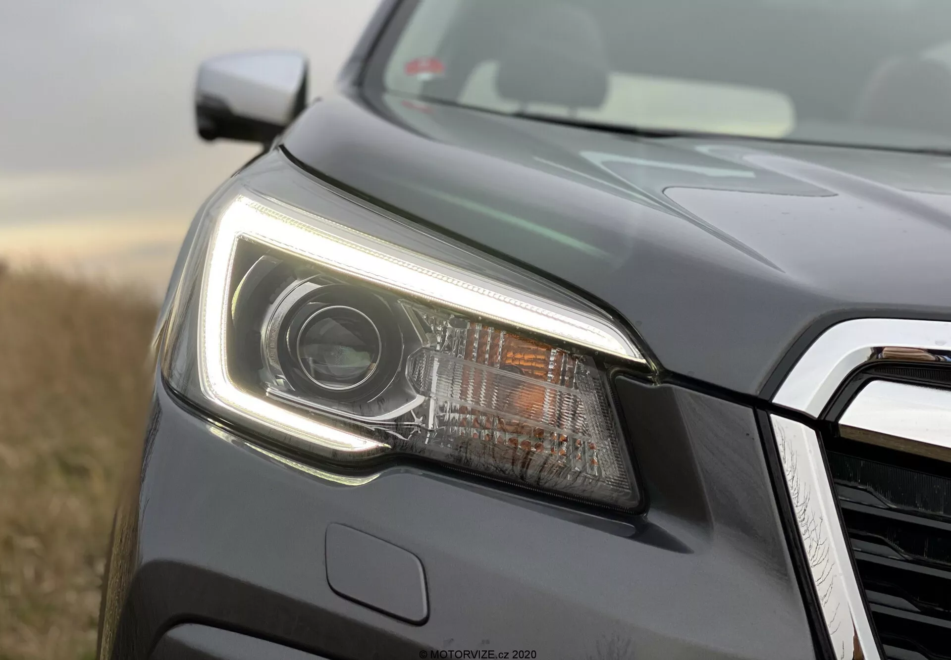 Primer plano del lado izquierdo delantero de un Subaru Forester 2020, centrado en el conjunto de faros modernos. La imagen, tomada desde un ángulo bajo, presenta un faro destacado con una lente de proyector y iluminación LED, que incluye luces diurnas nítidas. La carcasa del faro, elegante y con líneas angulares y forma aerodinámica. Se ve una parte de la rejilla frontal con un acabado cromado o metálico. El vehículo, pintado en gris oscuro metálico, está al aire libre con luz natural y un fondo vegetativo desenfocado, lo que resalta los detalles del faro y de la carrocería con enfoque selectivo.