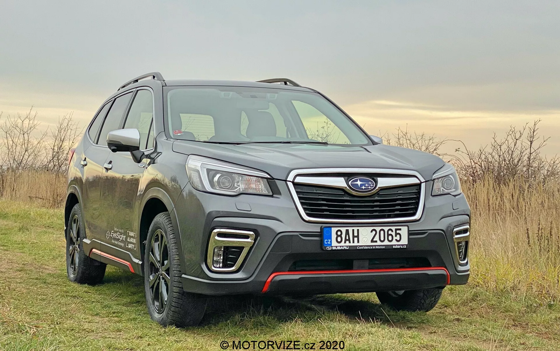 El Subaru Forester 2020 fotografiado, un SUV moderno capturado desde un ángulo de tres cuartos frontal. Posicionada ligeramente a la derecha del encuadre en un parche fuera de carretera con césped, bajo un cielo nebuloso. La imagen resalta características clave como la fascia delantera, la parrilla con una lama horizontal cromada y un módulo sensor, faros LED detallados, líneas contorneadas del capó y un perfil lateral del conductor. Llanta de aleación visible con un diseño de múltiples radios, espejos laterales con señales de giro integradas y líneas de carrocería a lo largo de las puertas sugieren un acabado de mayor categoría. El barro en el vehículo sugiere su uso fuera de la carretera y en caminos sin pavimentar. La suave luz diurna y el clima nublado aseguran una iluminación uniforme, enfatizando el diseño y las capacidades del coche.