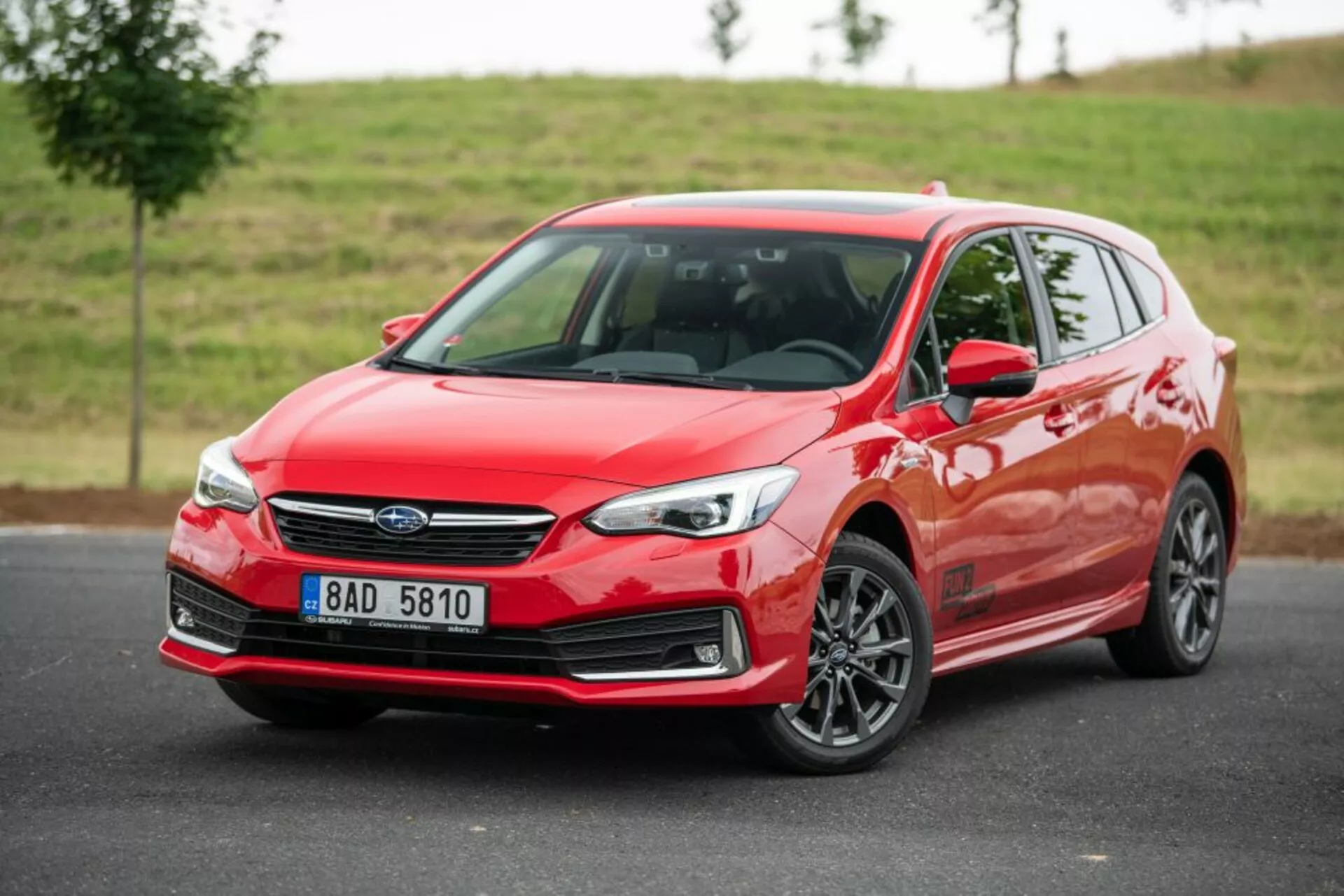 Subaru Impreza hatchback rojo brillante de 2018 con vista frontal de tres cuartos, mostrando sus elegantes faros LED, parrilla distintiva con emblema central, y llantas de aleación de múltiples radios contra un fondo natural nublado