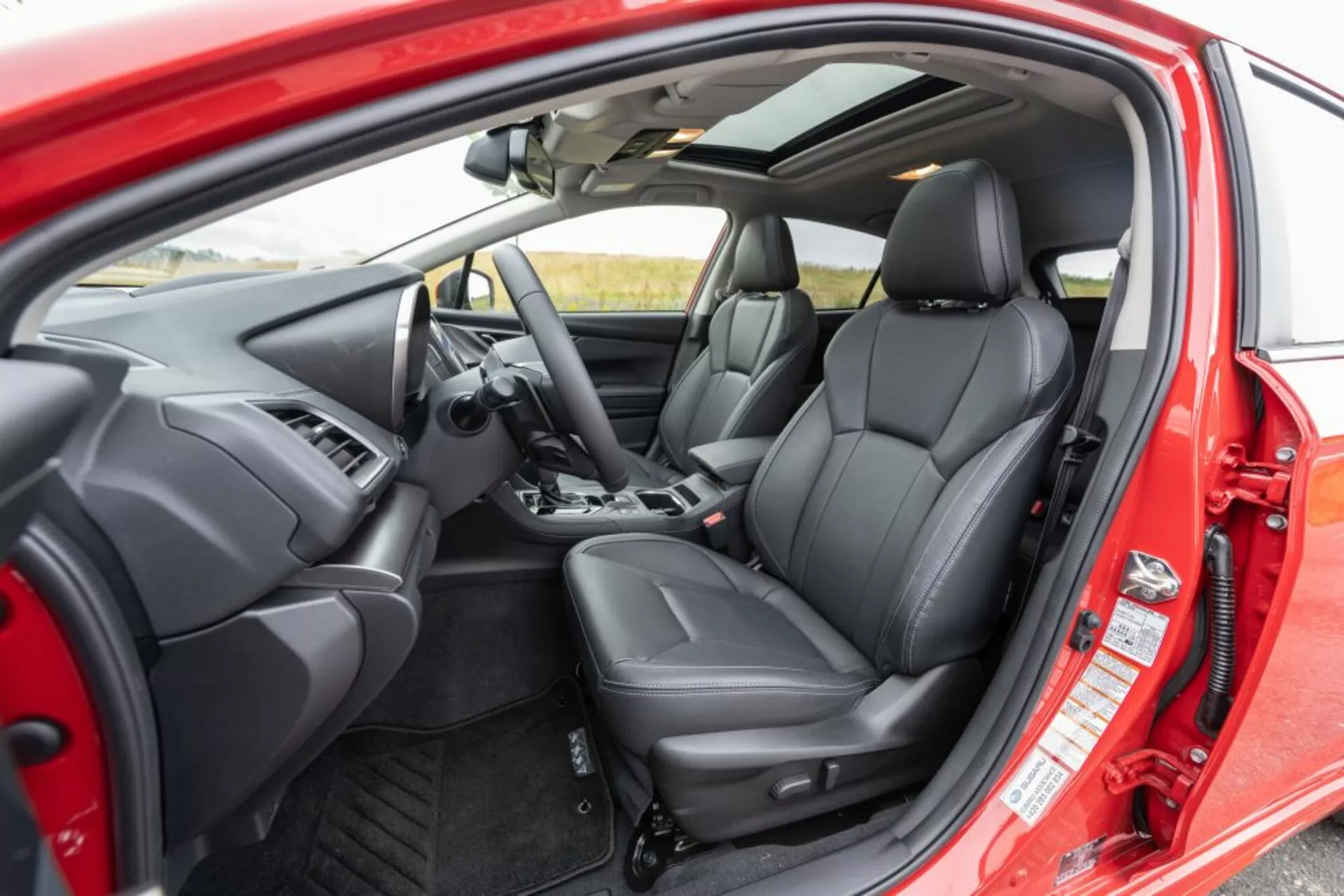 Vista interior de Subaru Impreza 2018 desde una perspectiva exterior mostrando el asiento del conductor, el tablero, el volante con botones de control, asientos delanteros ergonómicos de cuero negro y controles interiores de puerta a través de la puerta del conductor abierta