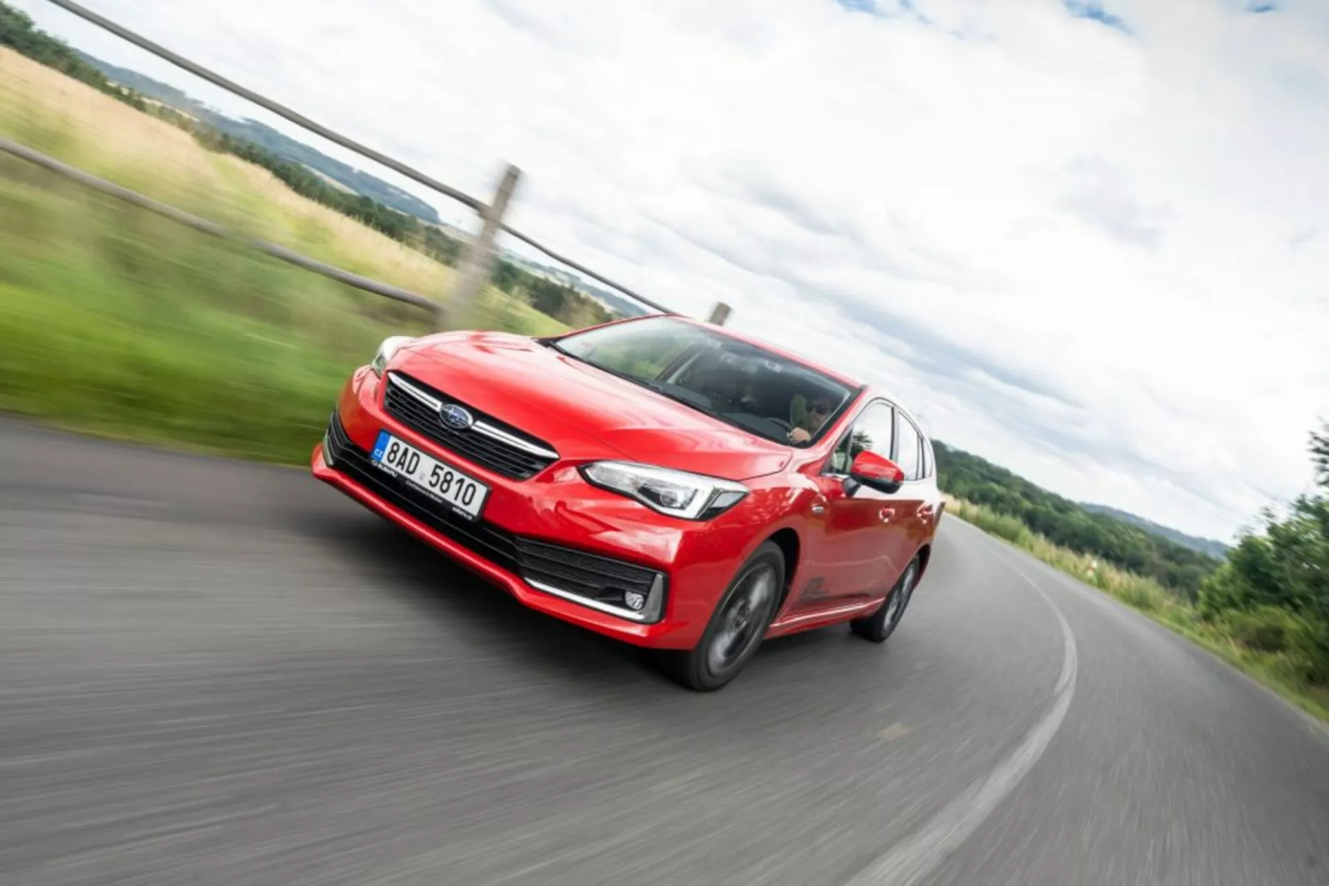 Imagen dinámica con desenfoque de movimiento de un Subaru Impreza rojo del 2018 en acción, vista frontal que muestra faro, parrilla y ruedas de aleación durante un giro a la izquierda en una carretera rural