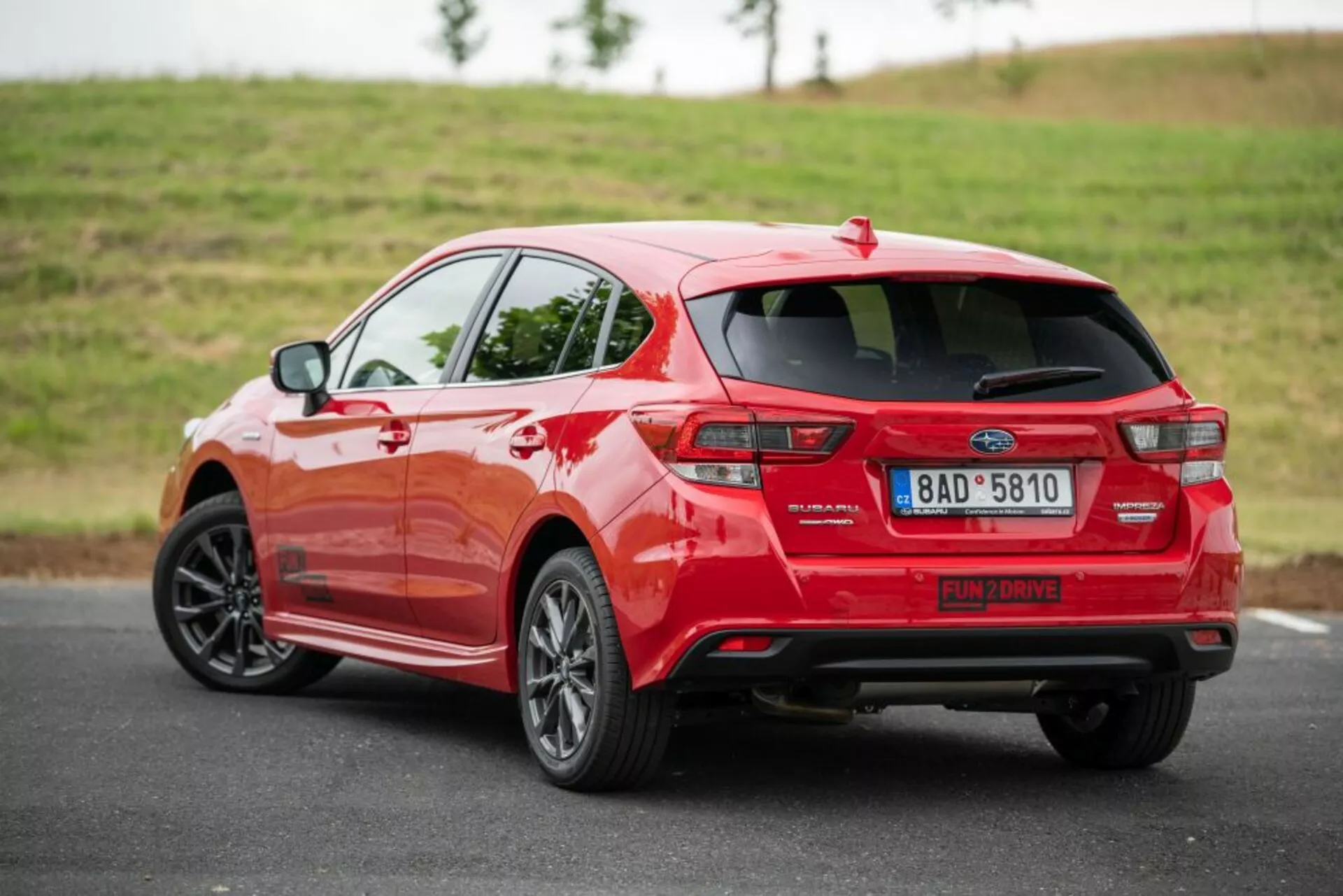 Hatchback rojo 2018 en vista trasera de tres cuartos en asfalto con una colina grassa de fondo, con spoiler montado en el techo, antena aleta de tiburón y sensores de aparcamiento visibles