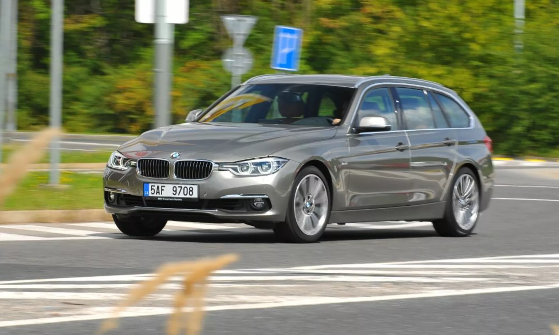 Vista dinámica de tres cuartos frontal de un BMW station wagon plateado en movimiento con la distintiva parrilla en forma de riñón y emblema, capturado desde una elevación leve que indica velocidad y movimiento, enfocándose en características de diseño y tecnología como faros angulares, ruedas de aleación y barras en el techo en un ambiente exterior con desenfoque de movimiento.