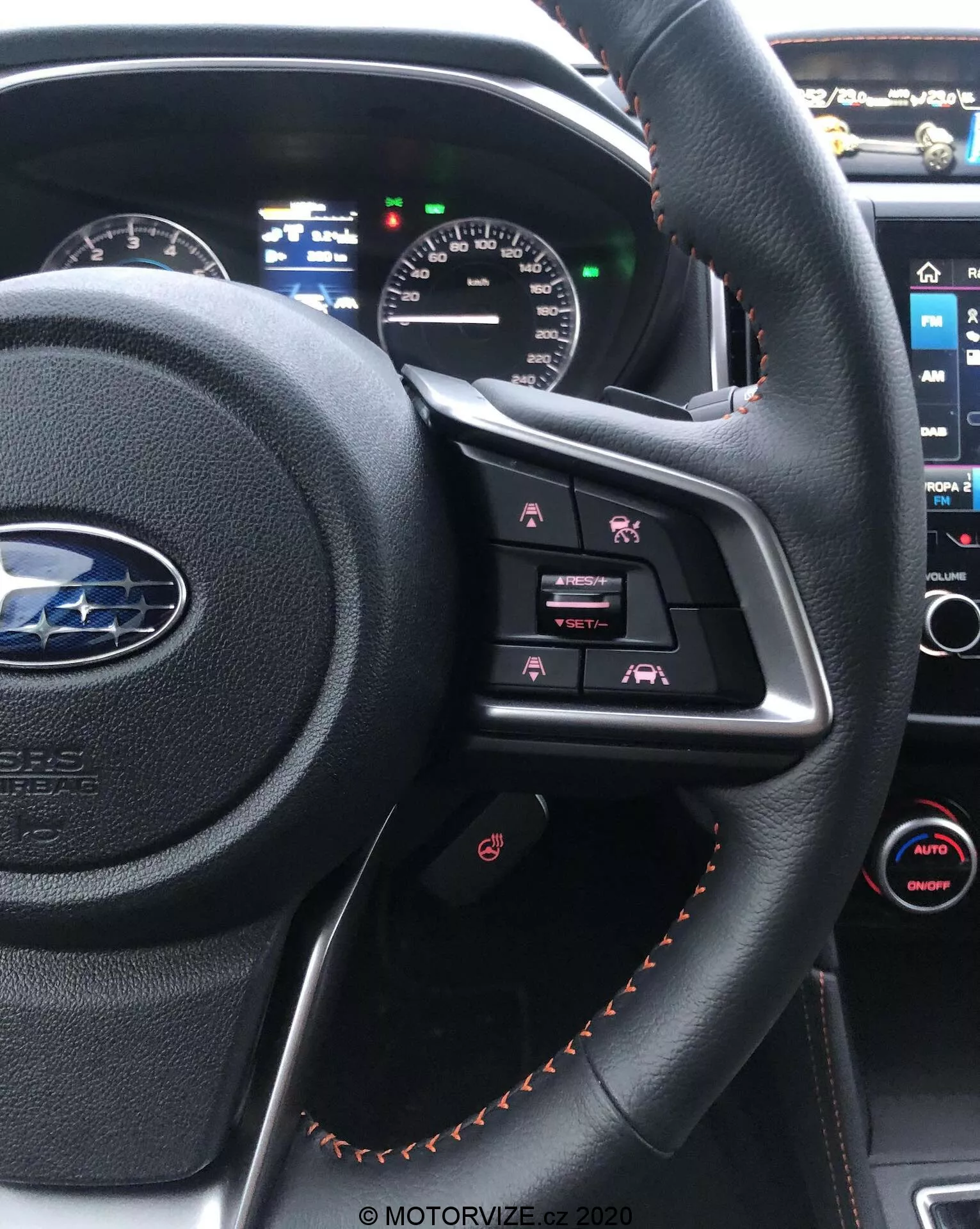 Vista interior de un Subaru XV 2018 desde el asiento del conductor, destacando el volante forrado en cuero negro con costuras en naranja, con botones de control multimedia y un panel de instrumentos parcialmente visible con pantallas analógicas y digitales. En el fondo se puede ver la consola central con su pantalla táctil de infoentretenimiento y el control climático automático de doble zona.