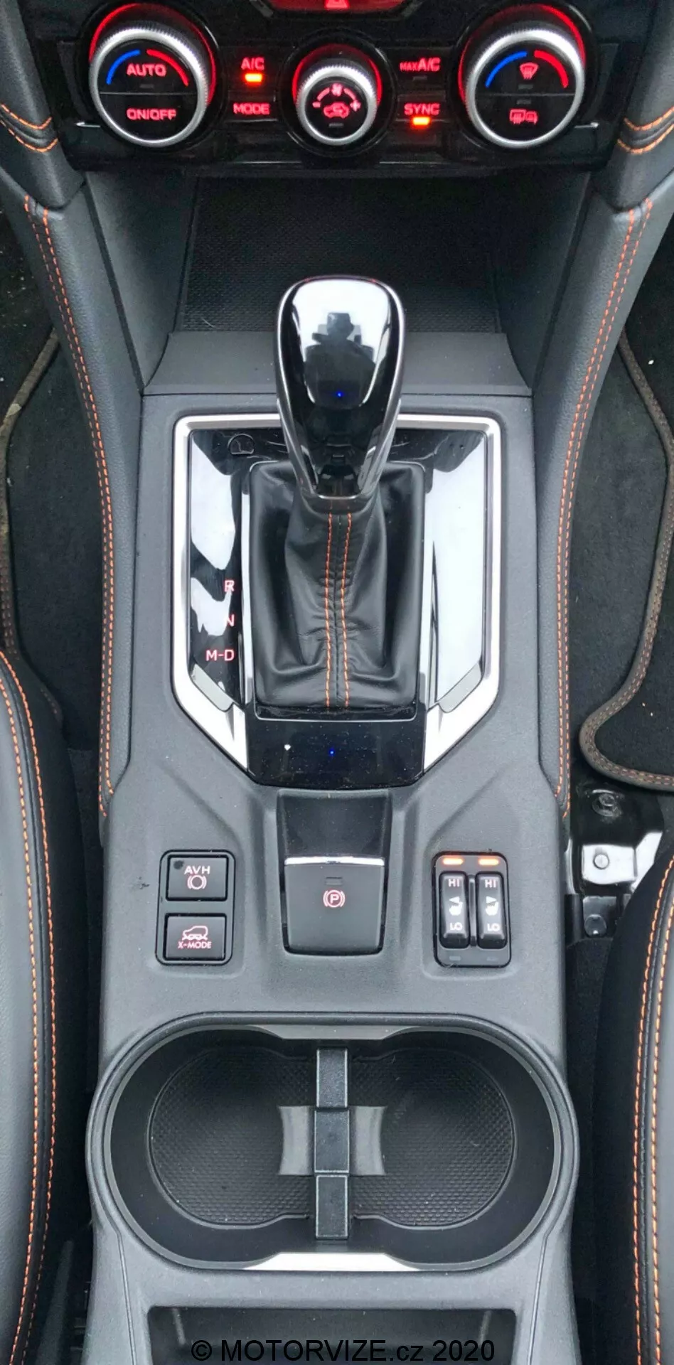 Vista vertical en primer plano de la consola central del Subaru XV 2018, que muestra la palanca de cambios con acabado metálico, selector de modos de conducción, varios botones de control incluyendo control de tracción, sensores de aparcamiento y configuraciones de suspensión, sistema de control climático de triple zona, detalles de costuras hechas a mano en cuero, portavasos doble, área de almacenamiento con puertos USB y marca de agua visible MOTORVIZE.cz.