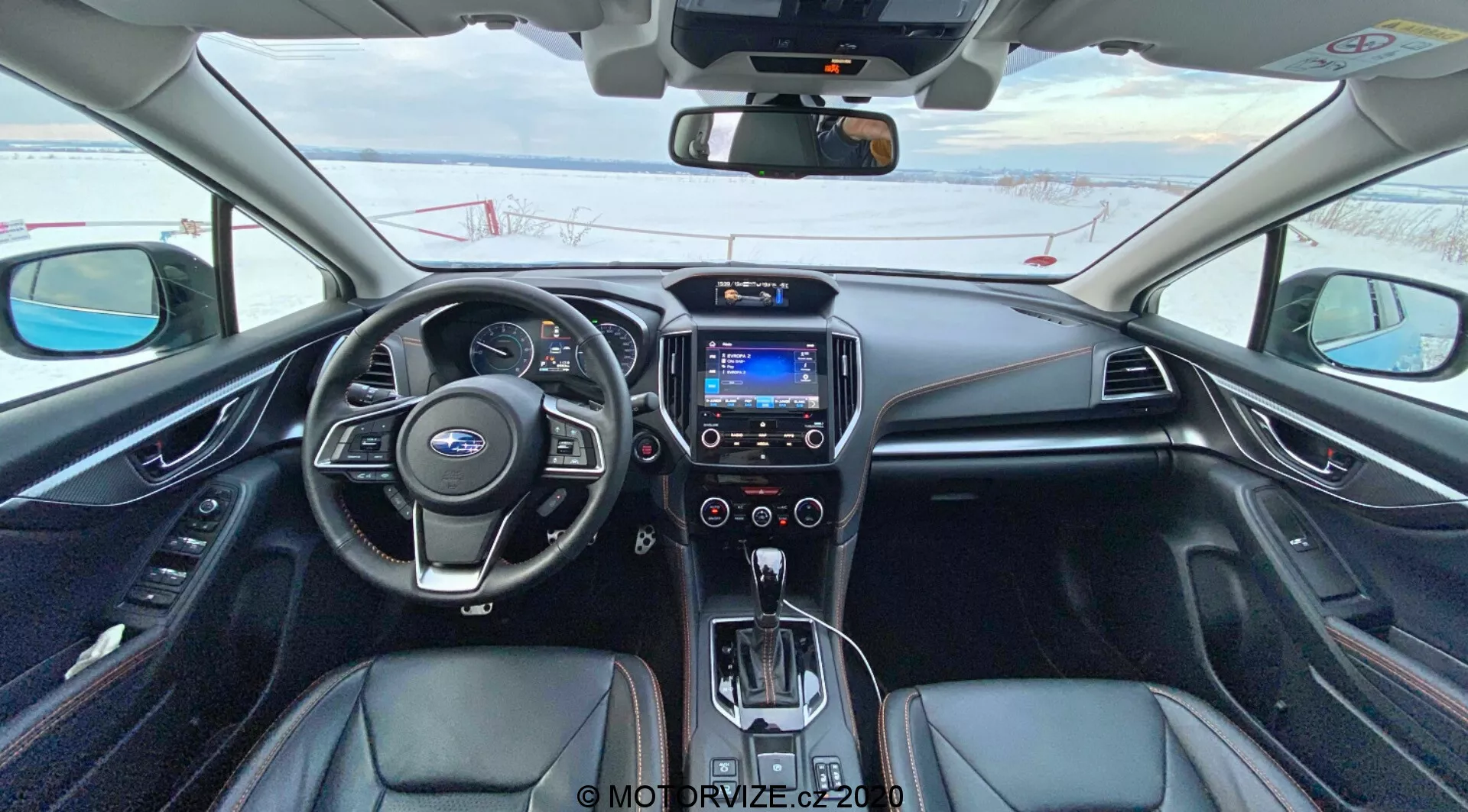 Vista interior de un Subaru XV 2018 (Subaru XV 1.6i Trend Lineartronic) desde el asiento del conductor mostrando el volante de cuero con controles integrados, panel de instrumentos analógico y digital parcialmente visible, pantalla de infoentretenimiento central con interfaz azul, perillas de control climático, palanca de cambios automática rodeada de botones operativos, tapicería negra con acentos plateados, controles de calefacción de asientos, y espejo retrovisor con pantalla digital. El coche está estacionado y hay nieve afuera, indicando un clima frío.
