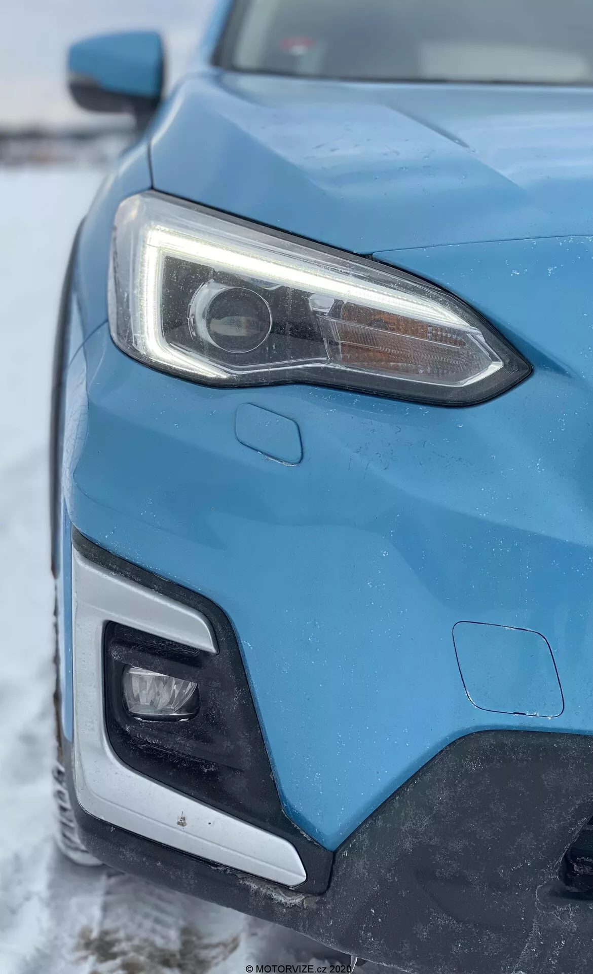 Vista de cerca del lado frontal izquierdo de un Subaru XV 1.6i Trend Lineartronic azul del 2018, capturada desde un ángulo oblicuo que se enfoca en el faro y una parte de la parrilla y el parachoques delantero. El faro izquierdo prominente presenta una lente de proyector indicativa de sistemas HID o LED y luces diurnas LED integradas. La imagen también muestra una parte del parachoques delantero que probablemente alberga iluminación adicional. La carrocería del automóvil exhibe líneas nítidas y formas aerodinámicas, con un telón de fondo invernal con una ligera capa de nieve.