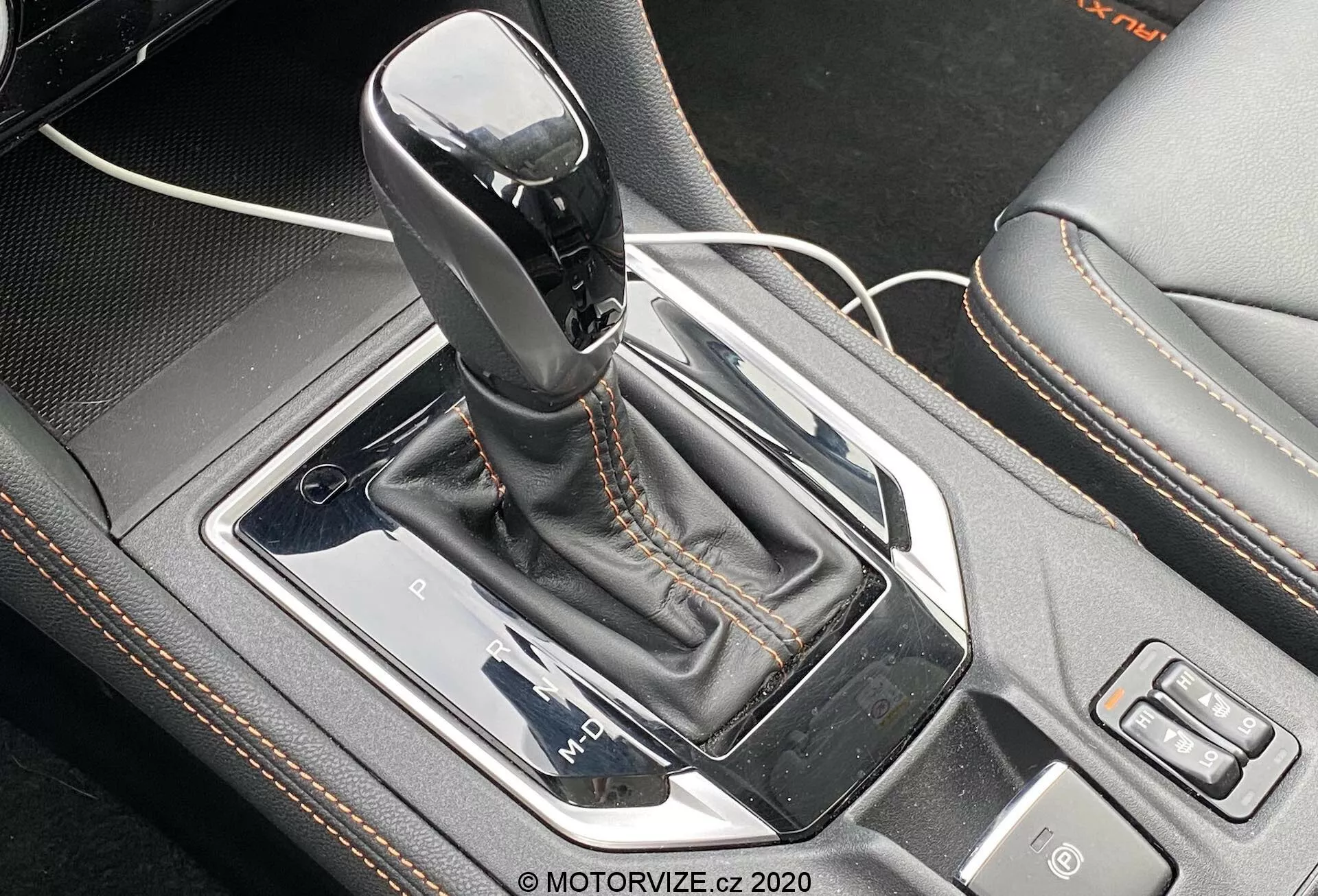 Vista de cerca de la palanca de cambios automática Lineartronic del Subaru XV 2018 1.6i Trend. La palanca de cambios, en la posición de estacionamiento y con una parte superior negra brillante con un acento plateado, está posicionada centralmente en la consola central del automóvil. Está rodeada por un panel plateado, rodeado por un bisel negro metálico de alto brillo con una funda de cuero sintético con costuras en contraste. A la izquierda, un cable USB indica un puerto cercano, mientras que a la derecha son visibles los botones para selección de modo de conducción y otra función. La perspectiva desde arriba y ligeramente por delante enfatiza los detalles de diseño intrincados y las características tecnológicas del área de la palanca de cambios.