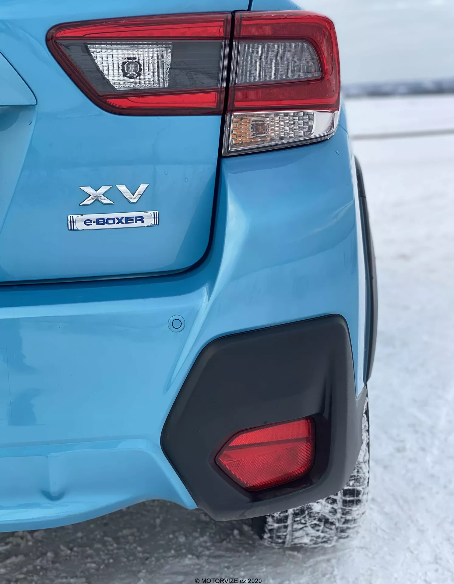 Un primer plano en ángulo trasero de un modelo Subaru XV 2018 azul, enfocándose en la esquina trasera izquierda desde la luz trasera hasta la rueda trasera. Las características clave incluyen un conjunto de luces traseras complejo, insignias 'XV' y 'e-BOXER' que indican tecnología híbrida, pintura metálica azul brillante con moldura de arco de rueda de plástico negro, un inserto de plástico negro en el parachoques trasero que refleja un suelo nevado que sugiere un clima frío, y un neumático trasero parcialmente visible con banda de rodadura cubierta de nieve. El enfoque está en la esquina trasera mientras que el fondo está ligeramente desenfocado debido a la profundidad de campo.