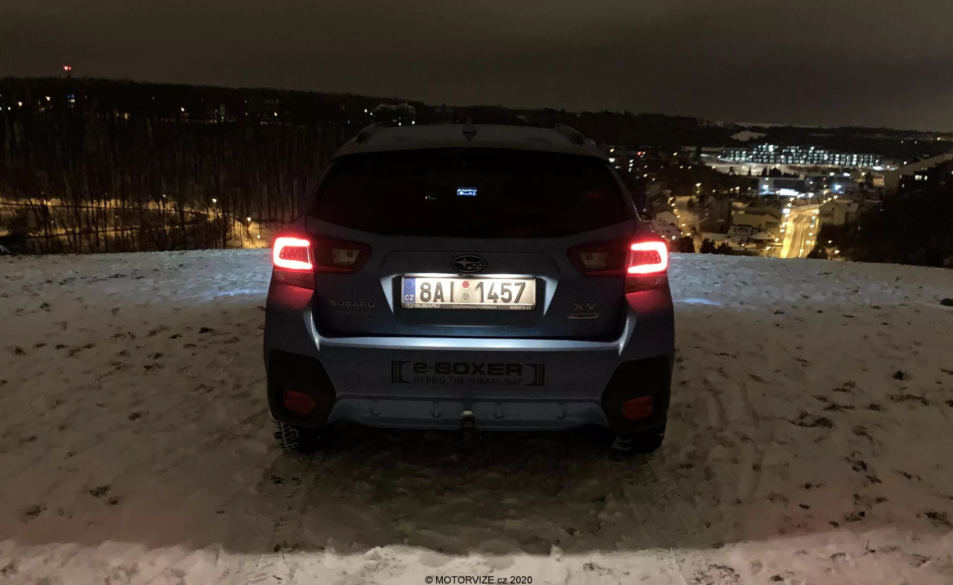 Vista trasera del Subaru XV 2018 (subaru-xv-1-6i-trend-lineartronic) capturada por la noche con las luces de la ciudad en el fondo. El coche está en un área elevada, mostrando las luces traseras iluminadas, el parabrisas trasero, el emblema del fabricante, la indicación del modelo e-BOXER, la matrícula, el parachoques trasero y la cámara o sensor de estacionamiento. Las superficies reflectantes y la nieve en el suelo sugieren condiciones invernales. El diseño moderno del vehículo incluye un pequeño alerón, un limpiaparabrisas trasero minimalista y un parachoques de dos tonos, destacando los elementos de iluminación automotriz y de diseño de marca.