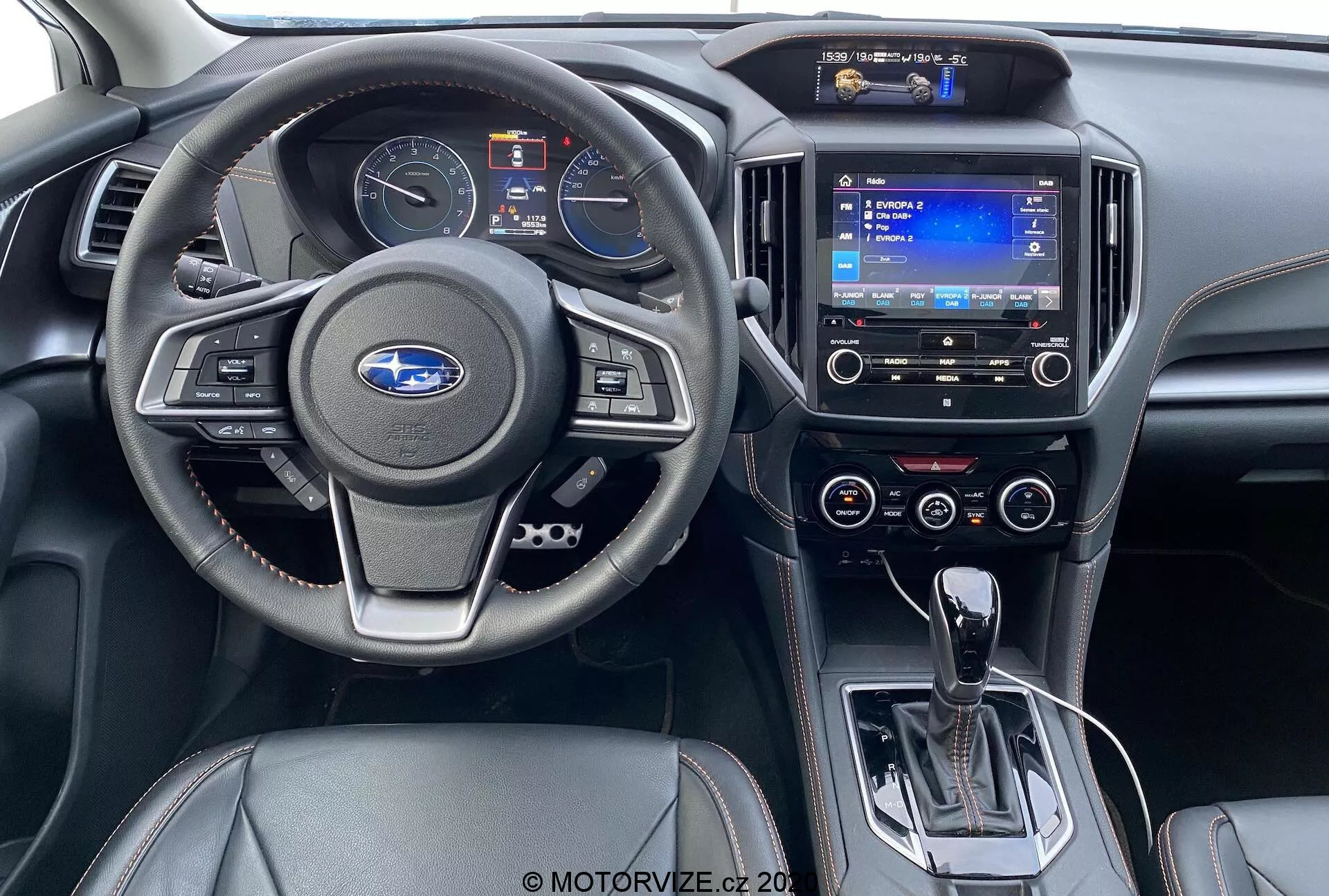 Vista interior desde el asiento trasero de un Subaru XV 1.6i Trend Lineartronic 2018. Presenta un volante moderno con botones de control de audio y crucero, un cuadro de instrumentos parcialmente visible, un sistema de infoentretenimiento con pantalla táctil y navegación, diales de control de clima de doble zona, palanca de cambios automática en posición de aparcamiento, interruptor de freno de mano electrónico, controles de calefacción de asientos y un interior de cuero negro de dos tonos con acentos plateados.