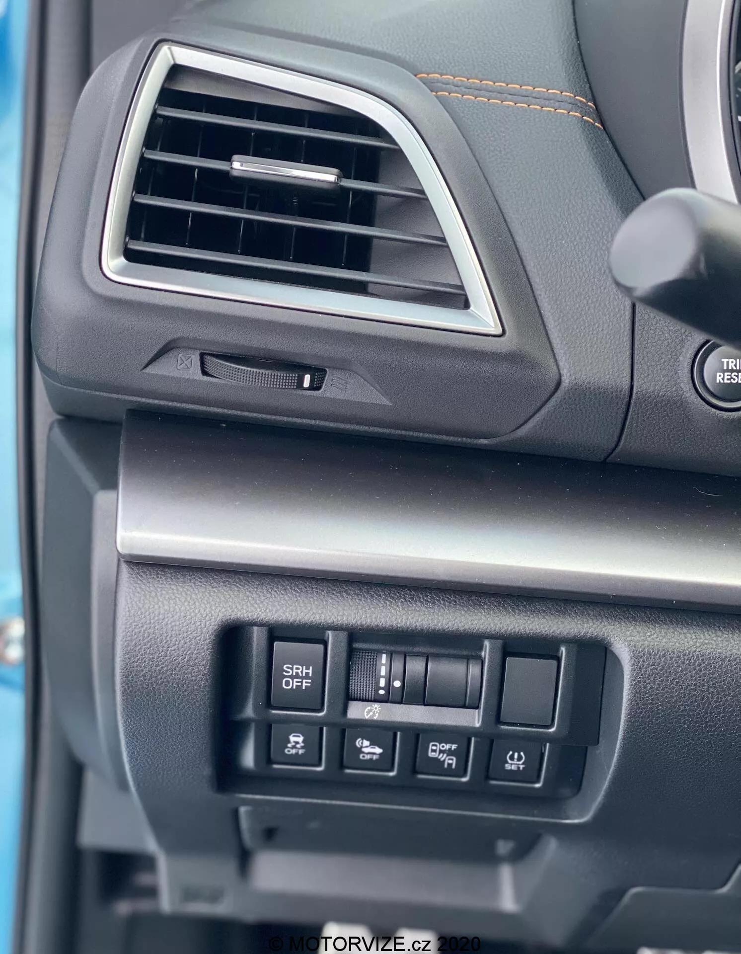 Vista de cerca del tablero del lado del conductor del Subaru XV 1.6i Trend Lineartronic 2018. La imagen se centra en una ventilación rectangular con bordes redondeados y un ajustador direccional, un volante parcialmente visible con botones de control de audio y un compartimento cerrado con una superficie suavemente texturizada debajo de la ventilación. También son visibles los botones de control para funciones como el Faros Responsivos a la Dirección (botón SRH OFF) y otros ajustes del vehículo. Los materiales del tablero parecen ser plásticos negros o grises oscuros con diferentes texturas, lo que indica durabilidad y calidad.