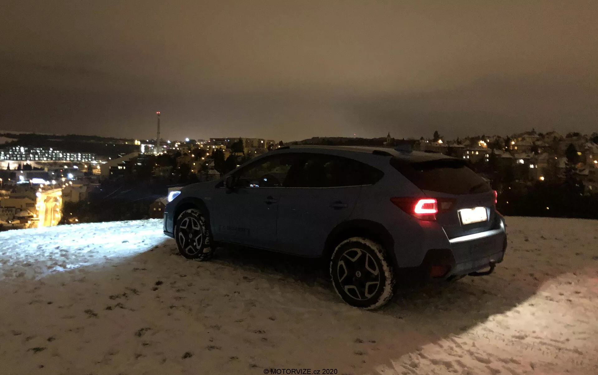 Subaru XV 1.6i Trend Lineartronic 2018 fotografiado de noche desde un ángulo trasero de tres cuartos, mostrando el lado derecho y la parte trasera, incluyendo las luces traseras, el parachoques trasero, la rueda trasera derecha, las puertas del pasajero y la rueda delantera derecha. El vehículo, probablemente de color azul oscuro o gris, está estacionado sobre una superficie cubierta de nieve con las luces de la ciudad visibles en la distancia. Las luces traseras indican que el coche podría estar encendido, y las distintivas ruedas más grandes indican una versión de nivel superior o deportiva. La escena enfatiza los componentes estéticos y el entorno ambiental sin visibilidad clara de características tecnológicas avanzadas.