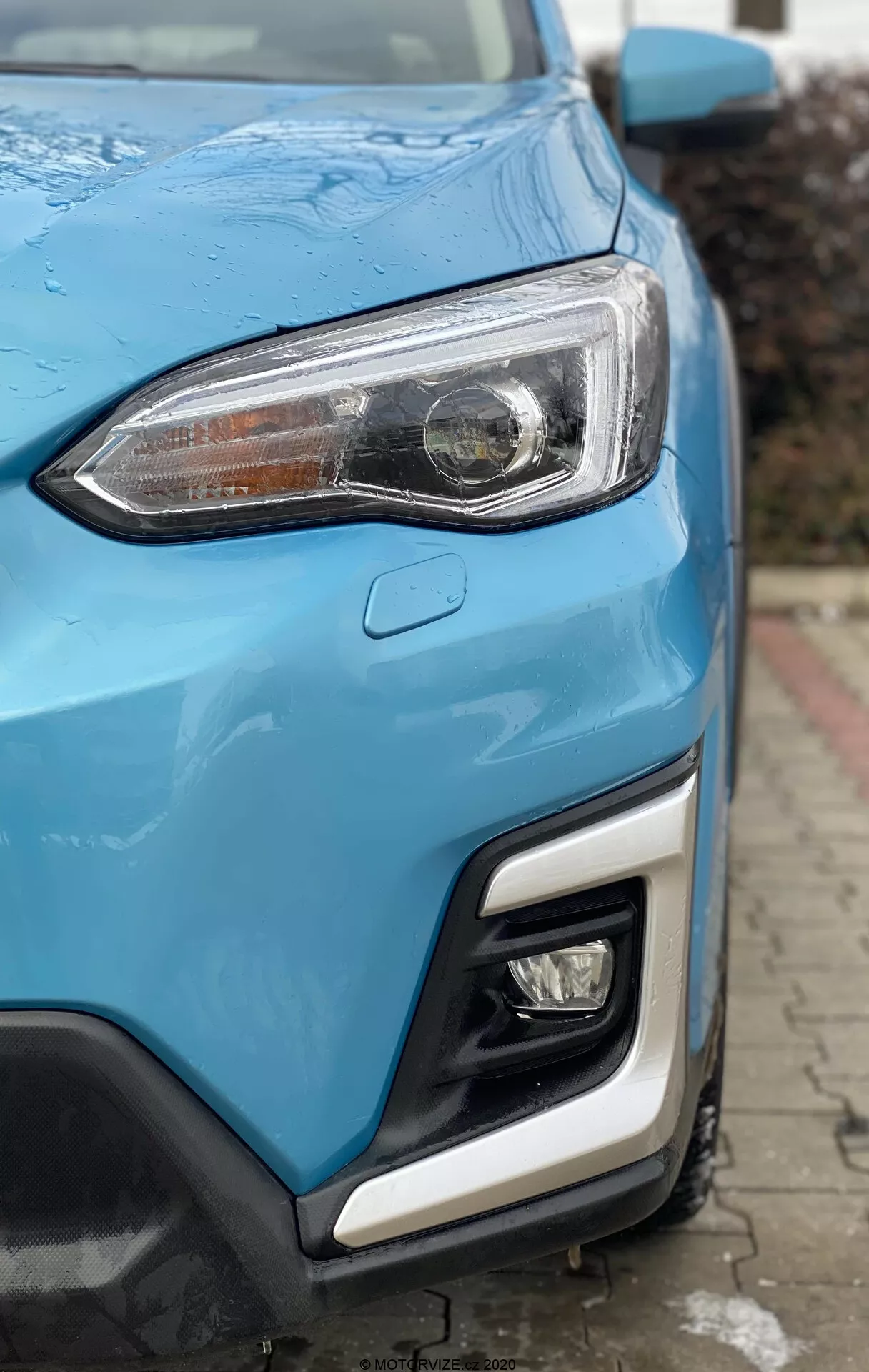 Vista de cerca de la esquina delantera de un Subaru XV 1.6i Trend Lineartronic 2018, mostrando un faro moderno con lente transparente y luces diurnas LED, parte de la parrilla y el parachoques en pintura azul vibrante. El parachoques cuenta con una luz antiniebla o secundaria y un sensor de estacionamiento. Los paneles del cuerpo elegantes y deportivos con gotas de agua sugieren lluvia reciente o limpieza, resaltando la curvatura del coche. Fondo urbano desenfocado.