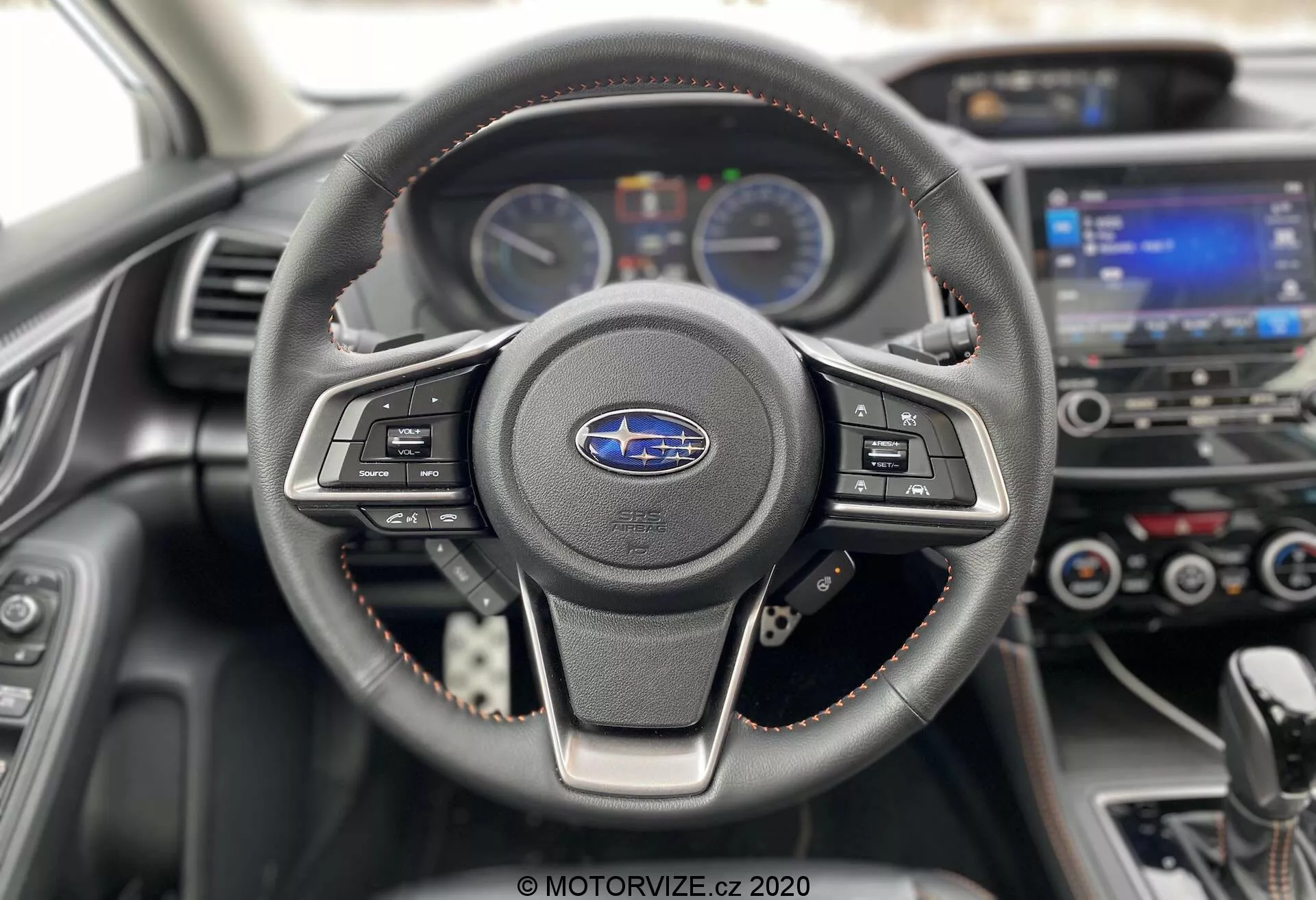 Perspectiva interior de un Subaru XV 2018 (Subaru-xv-1-6i-trend-lineartronic) enfocándose en el volante y el tablero de instrumentos. El volante de tres radios, envuelto en cuero con costuras visibles, cuenta con botones de control de multimedia y crucero. Detrás, el tablero muestra dos indicadores analógicos separados por una pantalla digital de información al conductor. En el fondo, ligeramente desenfocados, se ven el sistema de infoentretenimiento y los controles de clima. Las costuras rojas en el volante y el acolchado del tablero destacan un nivel de acabado deportivo o premium. Tomada desde el asiento del conductor, esta imagen moderna y bien mantenida enfatiza la tecnología y el diseño del automóvil.