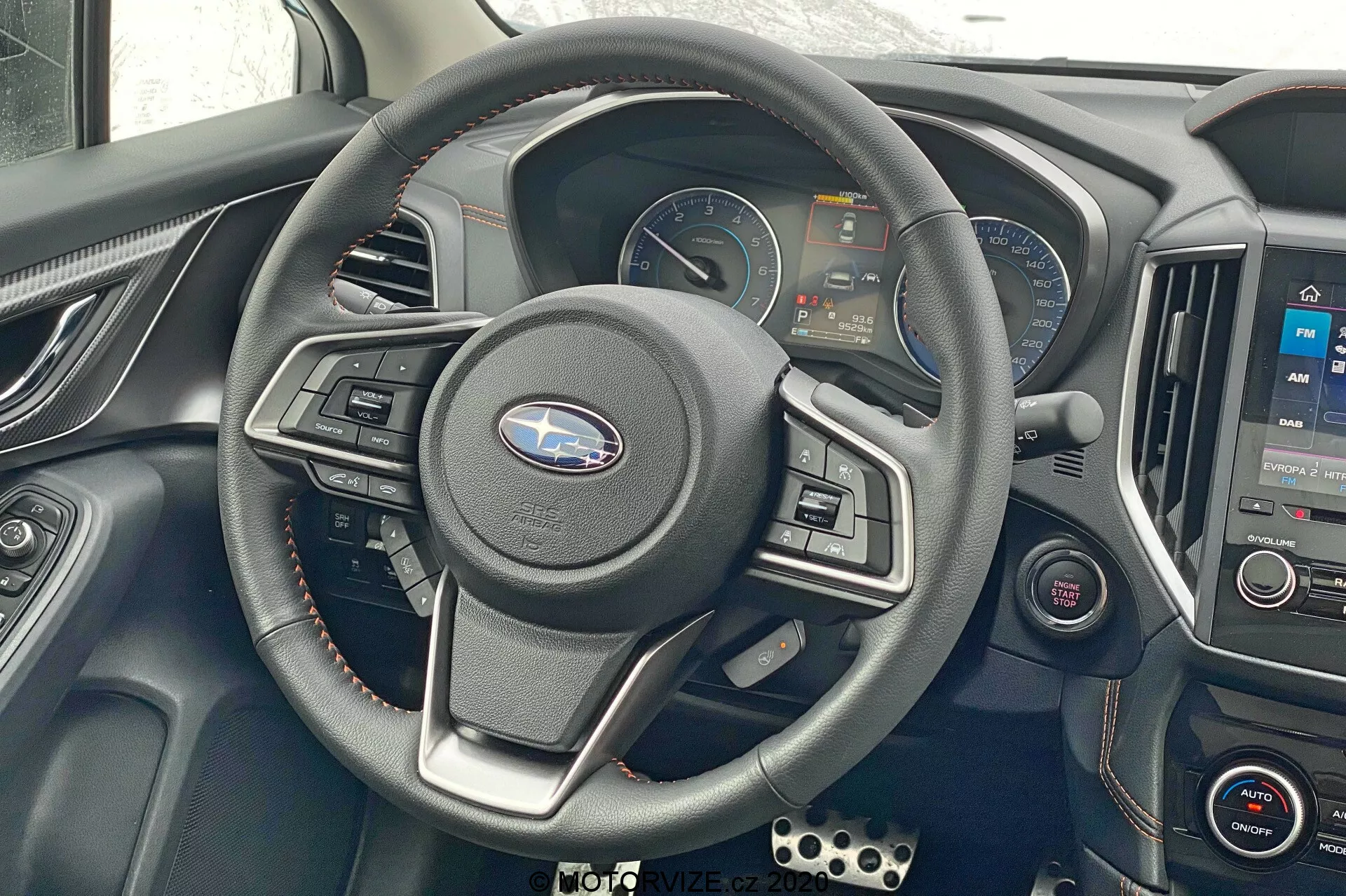 Toma interior del Subaru XV 2018, enfocada en el volante y el área del tablero. Volante de tres radios central con controles integrados, panel de instrumentos digital que muestra velocímetro, tacómetro y pantallas de información. Consola central con sistema de infoentretenimiento mostrando un mapa y controles de clima debajo. Detalles metálicos, volante revestido en cuero, detalles rojos en las ventilaciones. Botones de control adicionales a la izquierda, pedales estilo deportivo visibles en la parte inferior izquierda. Toma frontal, ligeramente hacia abajo mostrando controles y tecnología del conductor.