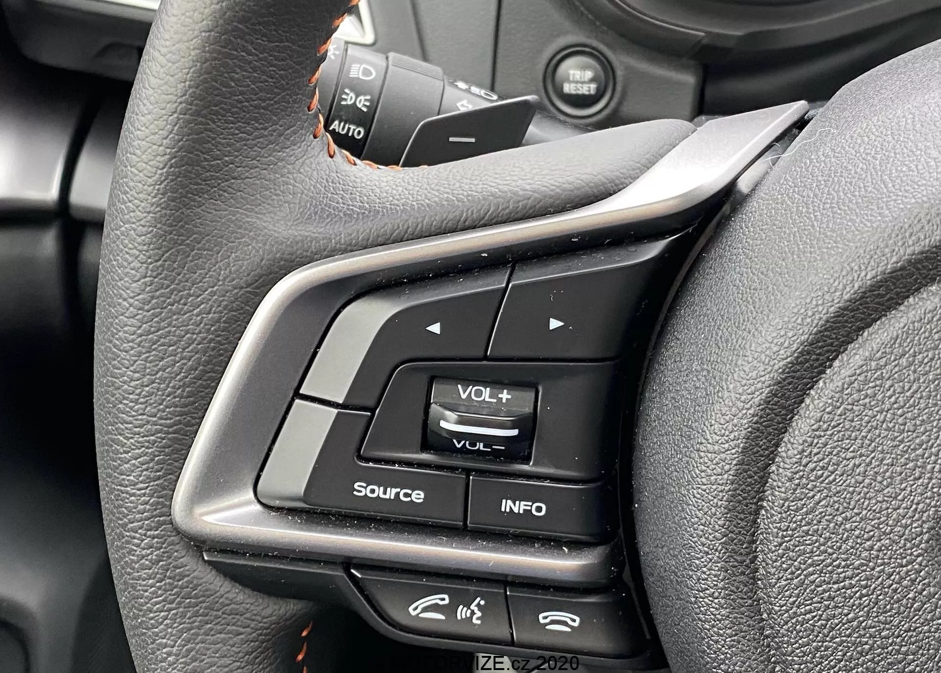 Vista en primer plano de los controles del volante del Subaru XV 2018 desde la perspectiva del asiento del conductor. La imagen resalta los botones de función múltiple en el radio izquierdo, incluyendo ajuste de volumen, botón INFO para configuraciones de pantalla, y un icono que parece un menú. Los interruptores del control de crucero están ligeramente fuera de foco arriba. Los controles están hechos de plástico y goma, con un volante envuelto en cuero que presenta un diseño ergonómico. Metadatos en la esquina inferior derecha leen 'IZE-EV2020'.