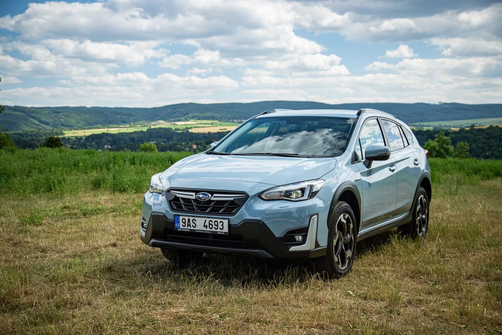 SUV crossover Subaru XV 2021 en un entorno natural al aire libre en un campo de hierba, mostrando parte frontal, capó, lado del conductor, vislumbre del techo, bajo luz diurna con cielo parcialmente nublado, destacando la rejilla con emblema, faros delanteros angulares afilados, luces de niebla integradas, ruedas de aleación modernas, espejos laterales con luces indicadoras, implicando avanzadas características de iluminación, seguridad y posible capacidad de tracción total.