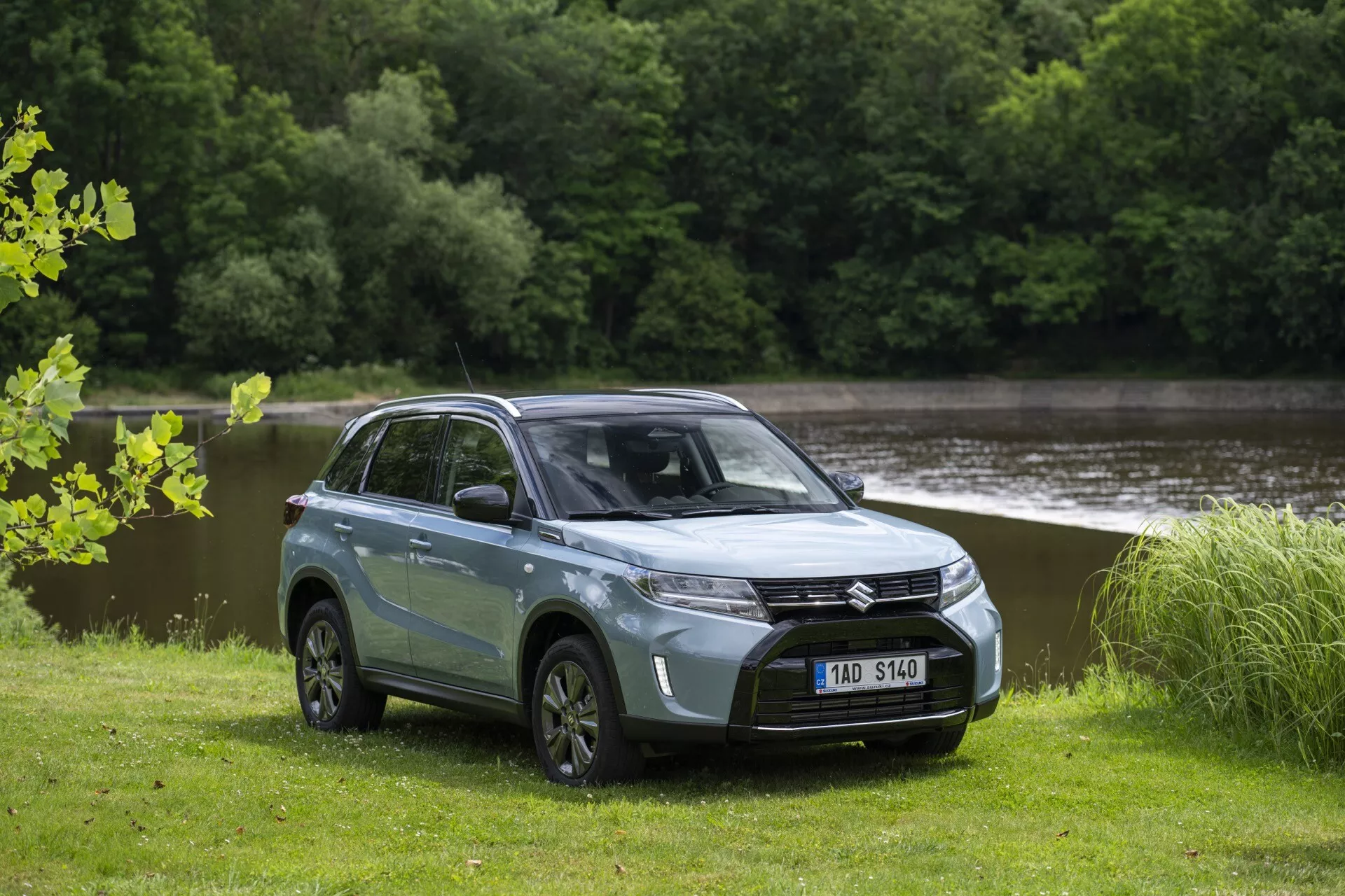 Fotografía de un SUV moderno Suzuki Vitara 2024 (2ª generación, facelift 2) en un cuerpo de dos tonos azul claro con techo negro, estacionado en un campo de hierba con agua y árboles en el fondo. El SUV, visto desde un ángulo de cuarto delantero que muestra el lado del conductor, presenta una parrilla delantera prominente con un emblema central, faros LED angulares elegantes, capó liso, ruedas de aleación multirrayos y una distancia al suelo notable. Las ventanas tintadas, los espejos laterales y las manijas de las puertas del color de la carrocería resaltan su diseño contemporáneo, mientras que los espejos laterales integran luces intermitentes LED.