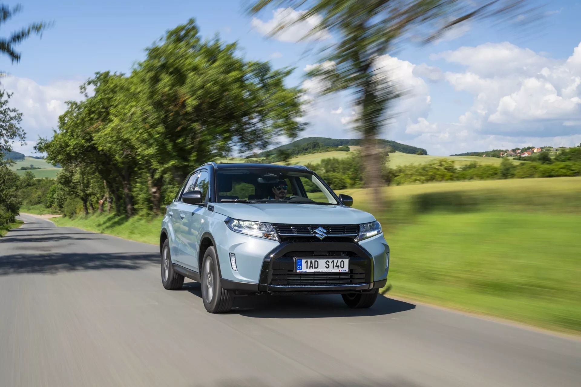 El Suzuki Vitara 2024 de 2ª Generación Facelift se captura en movimiento con un fondo borroso de árboles y cielo, enfatizando su velocidad. Esta vista en ángulo oblicuo frontal destaca el SUV moderno con una altura elevada, parrilla característica, luces de circulación diurna LED, faros antiniebla y tomas de aire. Los espejos laterales, el cuerpo aerodinámico y las ruedas de aleación en movimiento subrayan su diseño. Los sensores visibles sugieren sistemas avanzados de asistencia al conductor como control de crucero adaptativo y evitación de colisiones. La pintura metálica, el aspecto limpio y la matrícula clara pero anónima contribuyen a su estética. Posicionado en una carretera rural, muestra la versatilidad del vehículo para diversos desplazamientos.