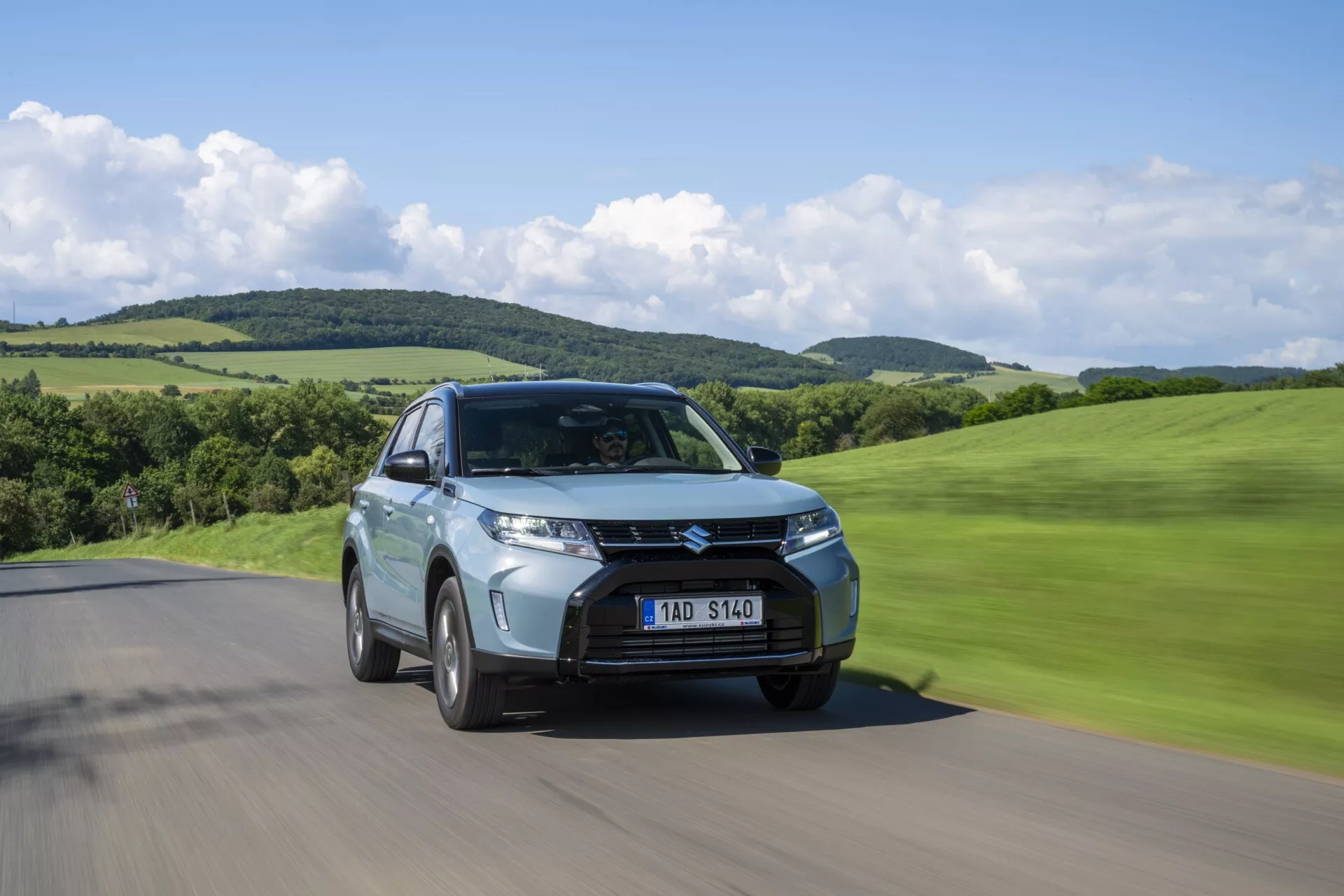 Toma en ángulo bajo de Suzuki Vitara 2024 de segunda generación facelift conduciendo en una carretera, vista de tres cuartos delanteros mostrando la parrilla, los faros y el desenfoque de movimiento en un paisaje verde y montañoso.