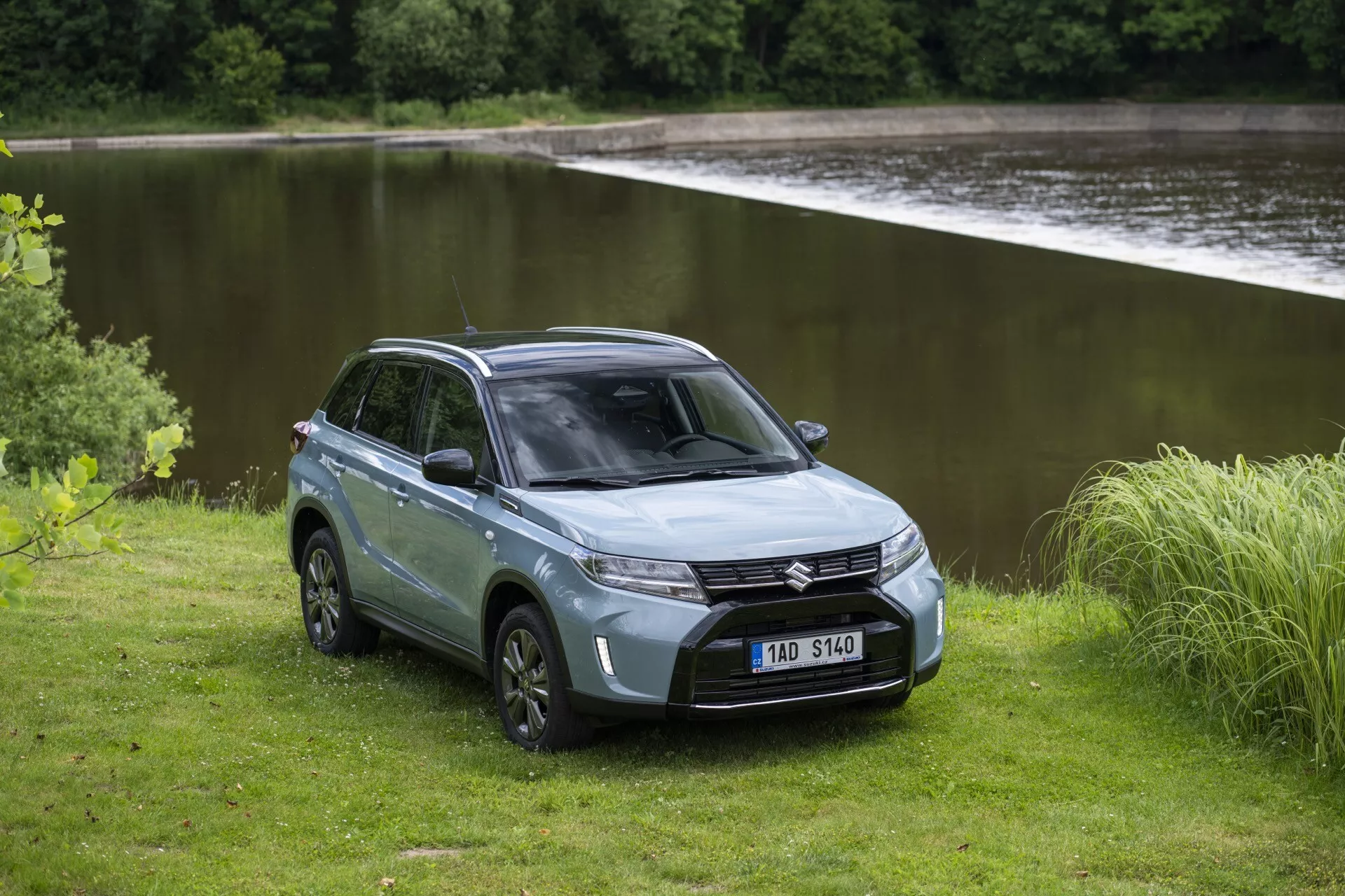 La imagen muestra el SUV Suzuki Vitara 2024 (segunda generación, segundo rediseño) en un entorno natural y sereno cerca de un cuerpo de agua. Capturado desde una vista frontal de tres cuartos, se pueden ver claramente los faros LED elegantes, la parrilla y el parachoques delantero. El capó esculpido y el parabrisas inclinado aportan un perfil aerodinámico. Un lado del SUV revela su diseño de cuatro puertas, área de carga trasera cerrada, llantas de aleación y neumáticos todoterreno robustos. El color plateado o gris apagado añade un toque refinado. El terreno natural y no pavimentado alrededor del SUV subraya sus capacidades todoterreno y su atractivo aventurero.