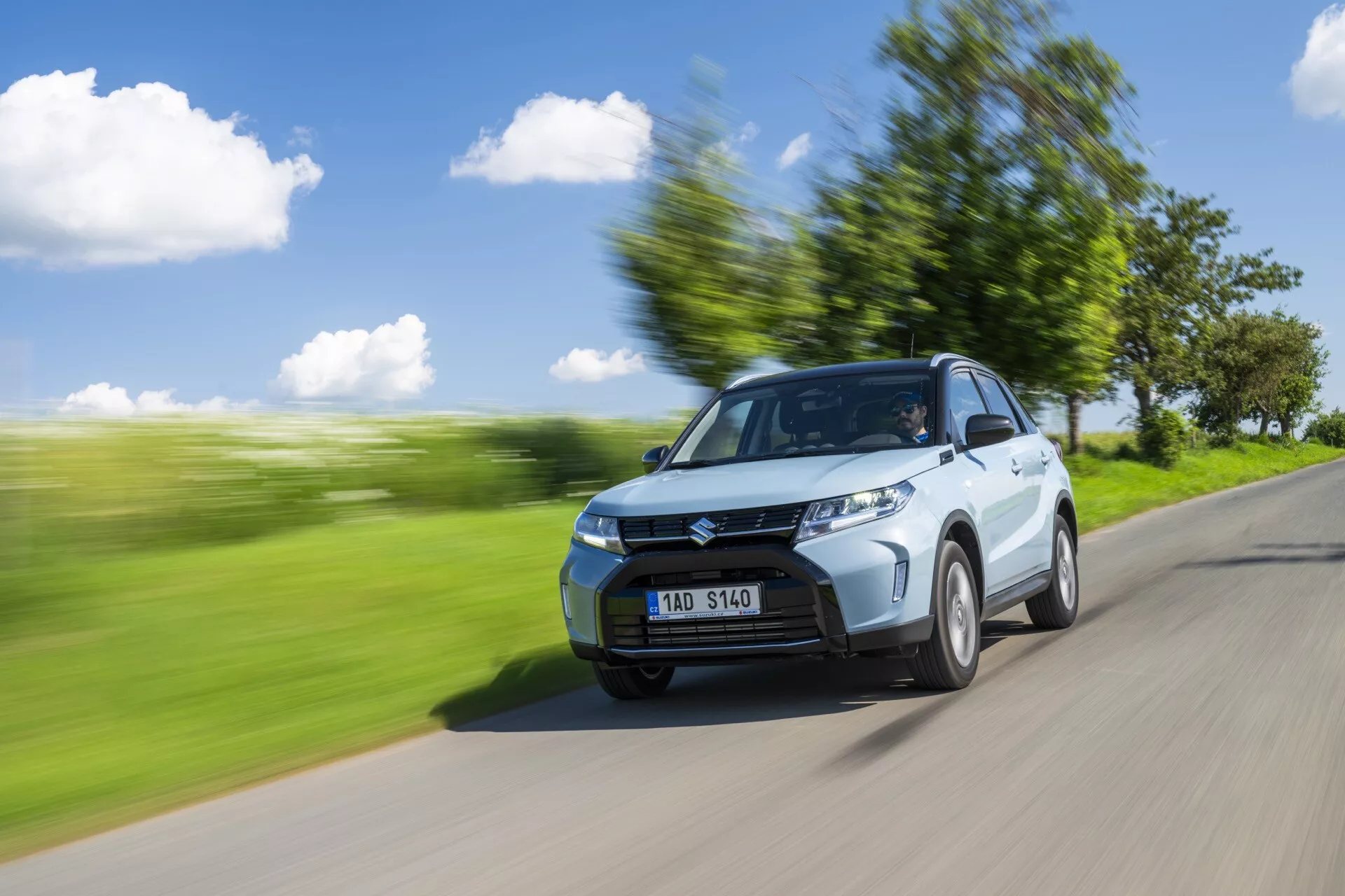 Toma dinámica del Suzuki Vitara 2024 (2da generación, facelift) capturada desde una vista frontal de tres cuartos. El auto circula por una carretera con un paisaje verde difuminado en el fondo, sugiriendo movimiento. Las características visibles incluyen el capó, parrilla, faros delanteros, parachoques frontal y una rueda delantera ligeramente girada. El costado del conductor, incluyendo la puerta y el retrovisor, es prominente. El desenfoque de movimiento y las rayas a lo largo del entorno mejoran la sensación de velocidad mientras el auto permanece enfocado. Se observan líneas de contorno, cortes laterales y el diseño de las ruedas. Condiciones de luz brillante con cielos despejados y sombras sugieren luz solar desde la parte superior izquierda. Se puede ver a una persona a través del parabrisas, probablemente el conductor.