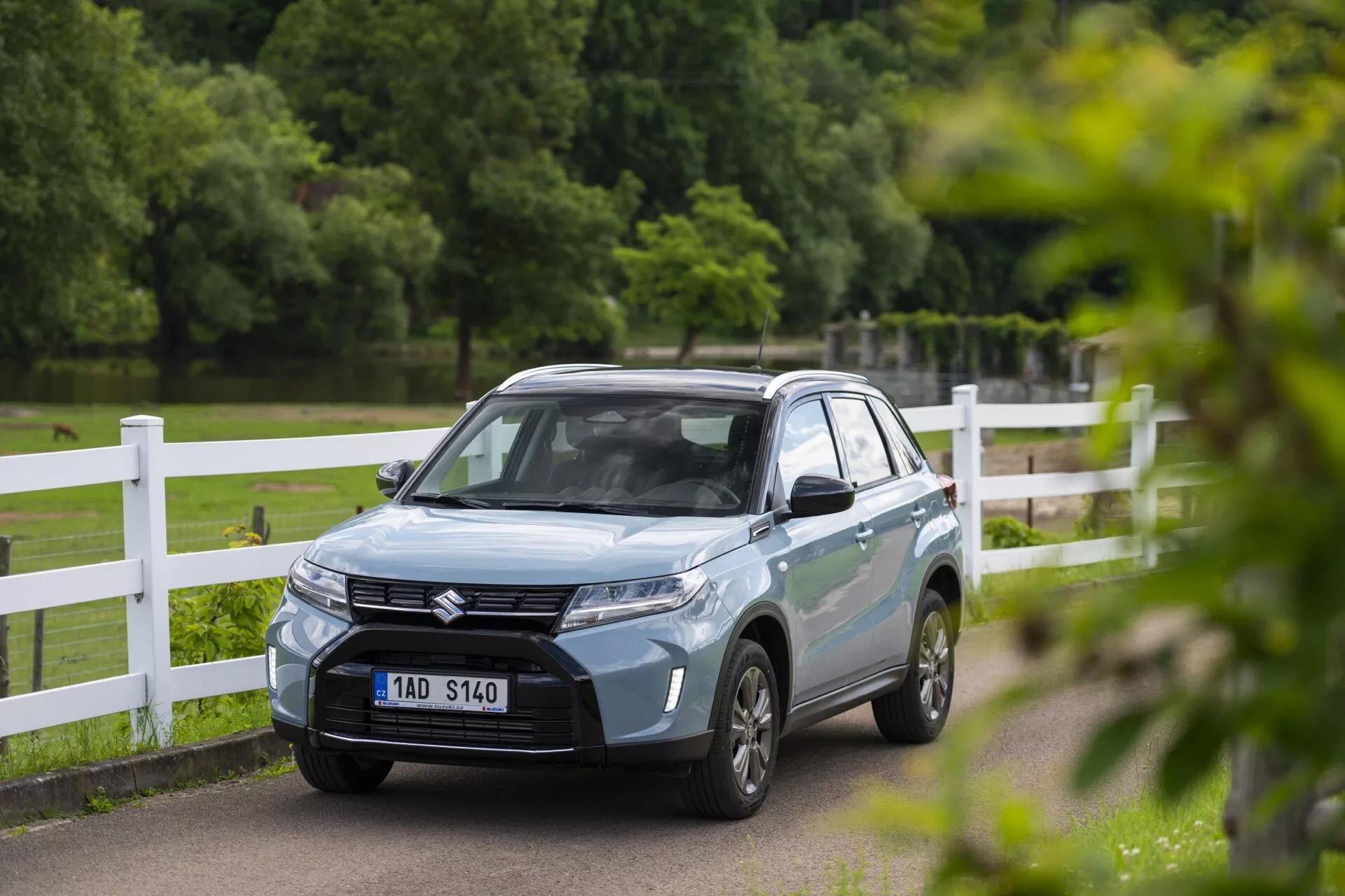 SUV Suzuki Vitara 2024 azul claro en un entorno de parque o campo, visto desde un ángulo delantero de tres cuartos que muestra su sofisticada parrilla con el emblema, faros LED afilados, líneas aerodinámicas en el capó y ruedas de aleación de varios radios. La matrícula delantera del vehículo está borrosa y los espejos retrovisores podrían tener señales de giro integradas. La foto captura el coche con poca profundidad de campo, enfatizando su diseño moderno mientras el fondo permanece ligeramente desenfocado.