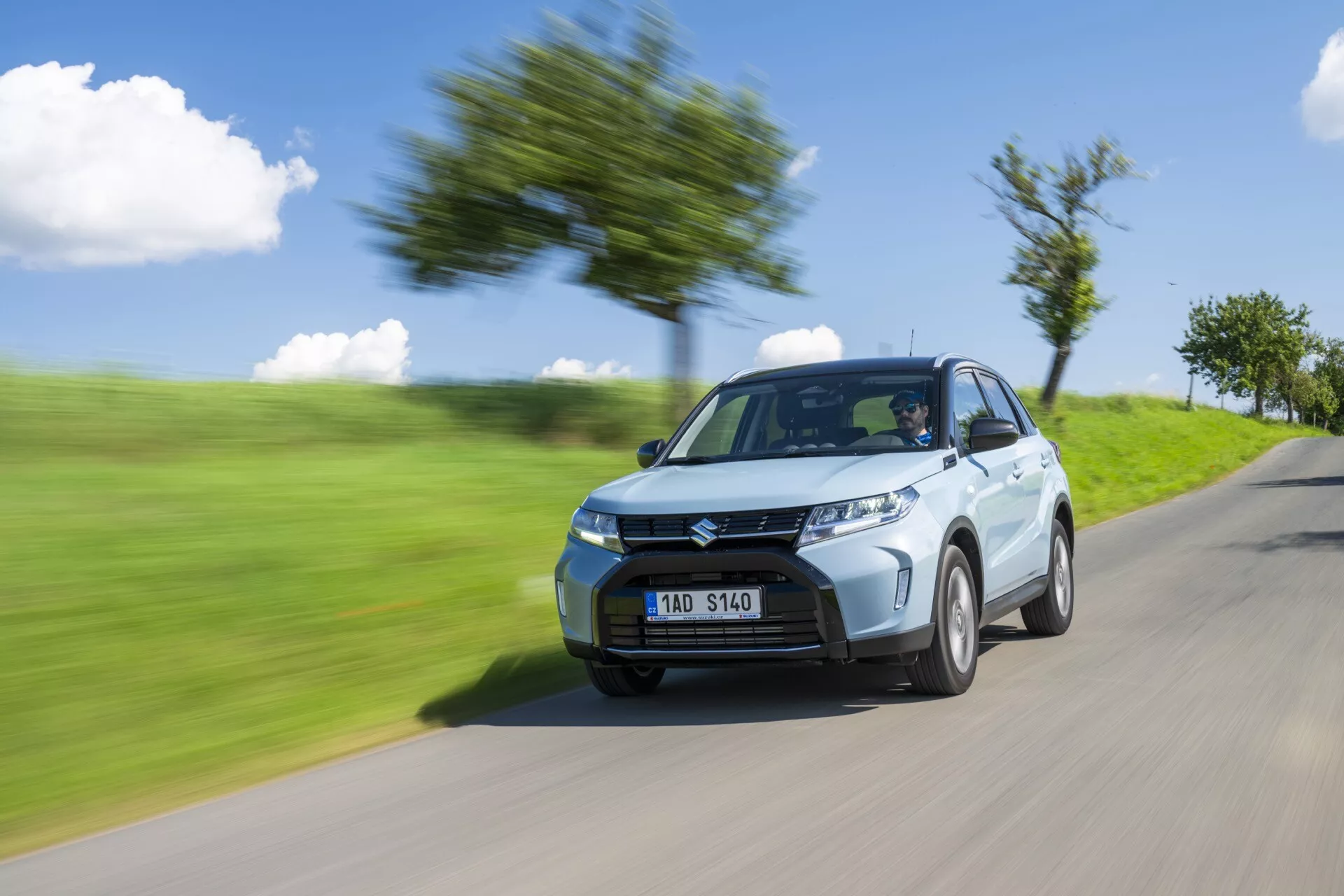 Toma dinámica del Suzuki Vitara 2024 (segunda generación facelift), mostrando su diseño moderno y velocidad. Capturado desde una perspectiva de cuarto delantero izquierdo, la imagen destaca los faros nítidos, la parrilla frontal prominente y las llantas de aleación estándar. El efecto de desenfoque de movimiento en el fondo y las ruedas acentúa el rápido movimiento del coche, visto en una carretera borrosa con árboles y hierba.