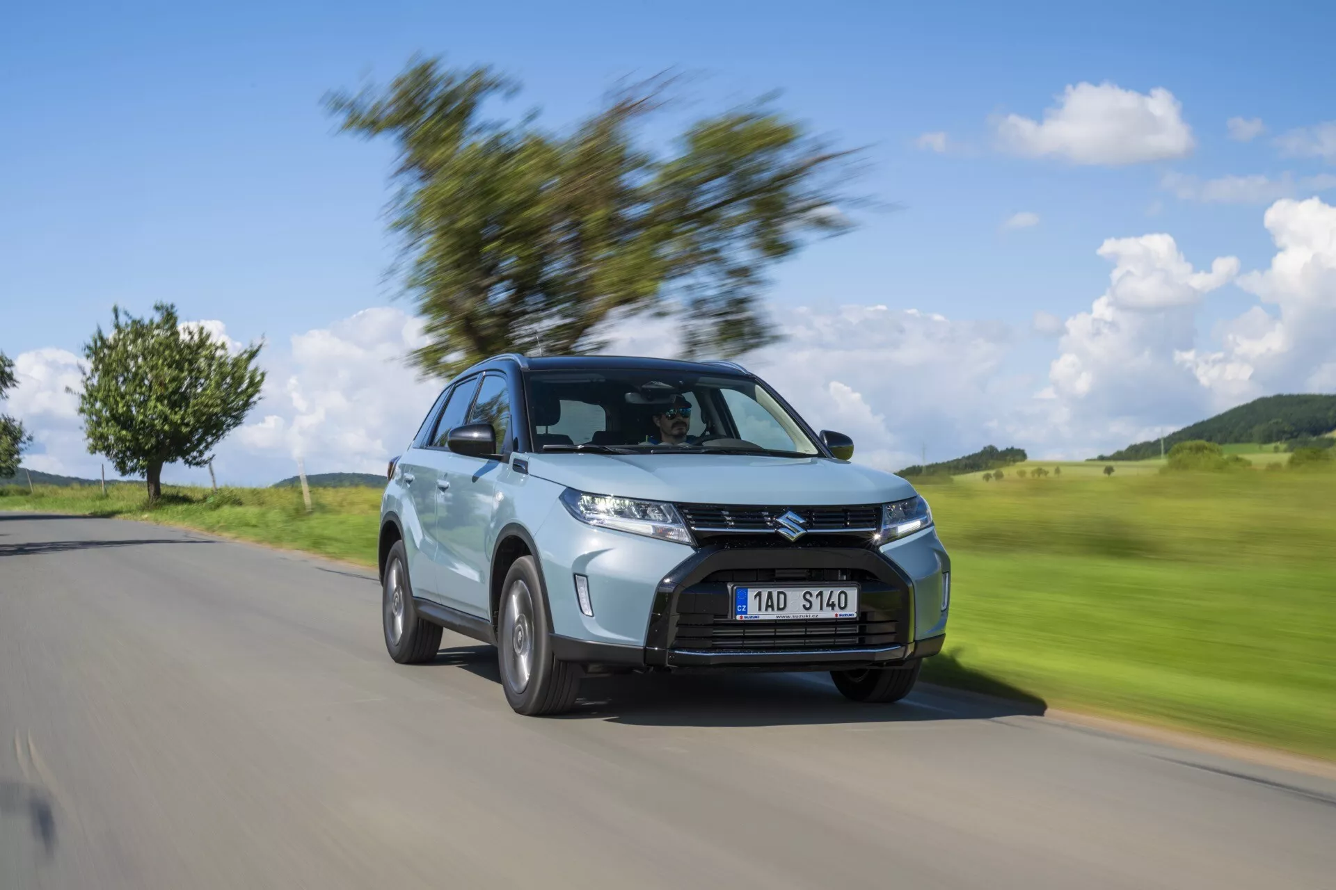 Imagen dinámica del Suzuki Vitara 2024 (2ª generación, facelift 2) en movimiento, mostrando el ángulo de tres cuartos delantero. El coche está enfocado con nitidez, mientras que el fondo está difuminado por movimiento, destacando sus faros, parrilla prominente, llantas de aleación, carrocería SUV compacta, pintura metálica y diseño aerodinámico. Ideal para exposiciones automotrices y uso promocional.
